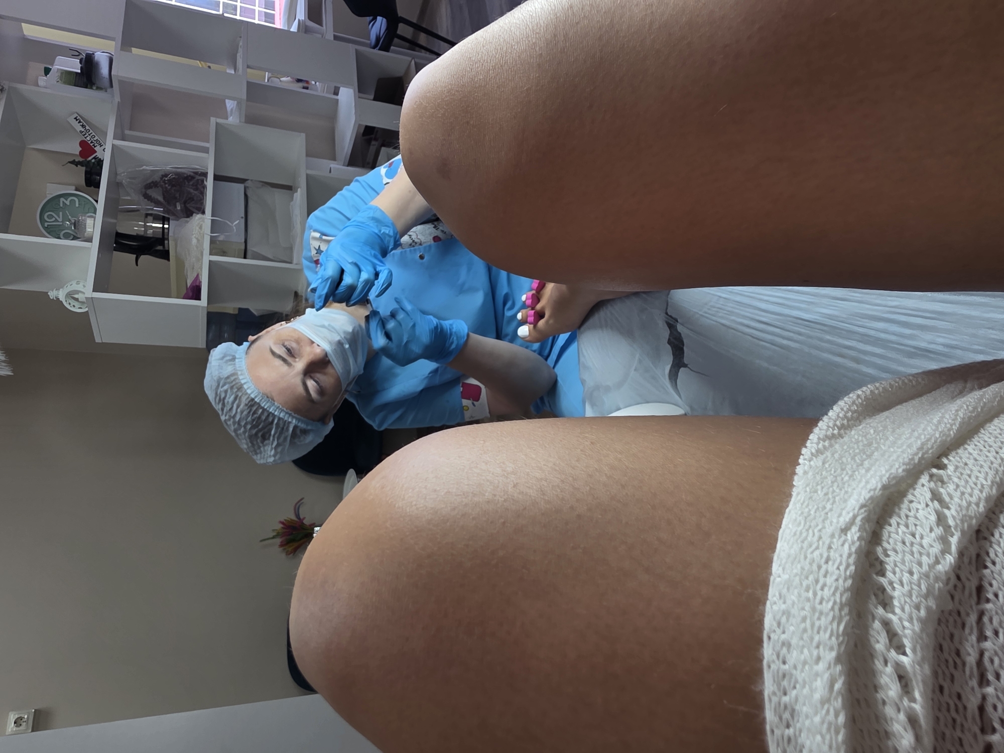 The image shows a close-up, point-of-view shot of a pedicure in progress at a salon in Sochi, Russia. The subject's legs, with one foot visible and adorned with pink toe separators, are in the foreground. A person, likely a nail technician, is working on the subject's toes. They are wearing a blue scrub suit, a hairnet, a face mask, and blue disposable gloves. The technician's hands are focused on the subject's toes, with tools and nail polish visible. In the background, a white shelving unit holds various items, including a clock, decorative objects, and bottles. The overall environment appears to be a clean and professional nail salon.