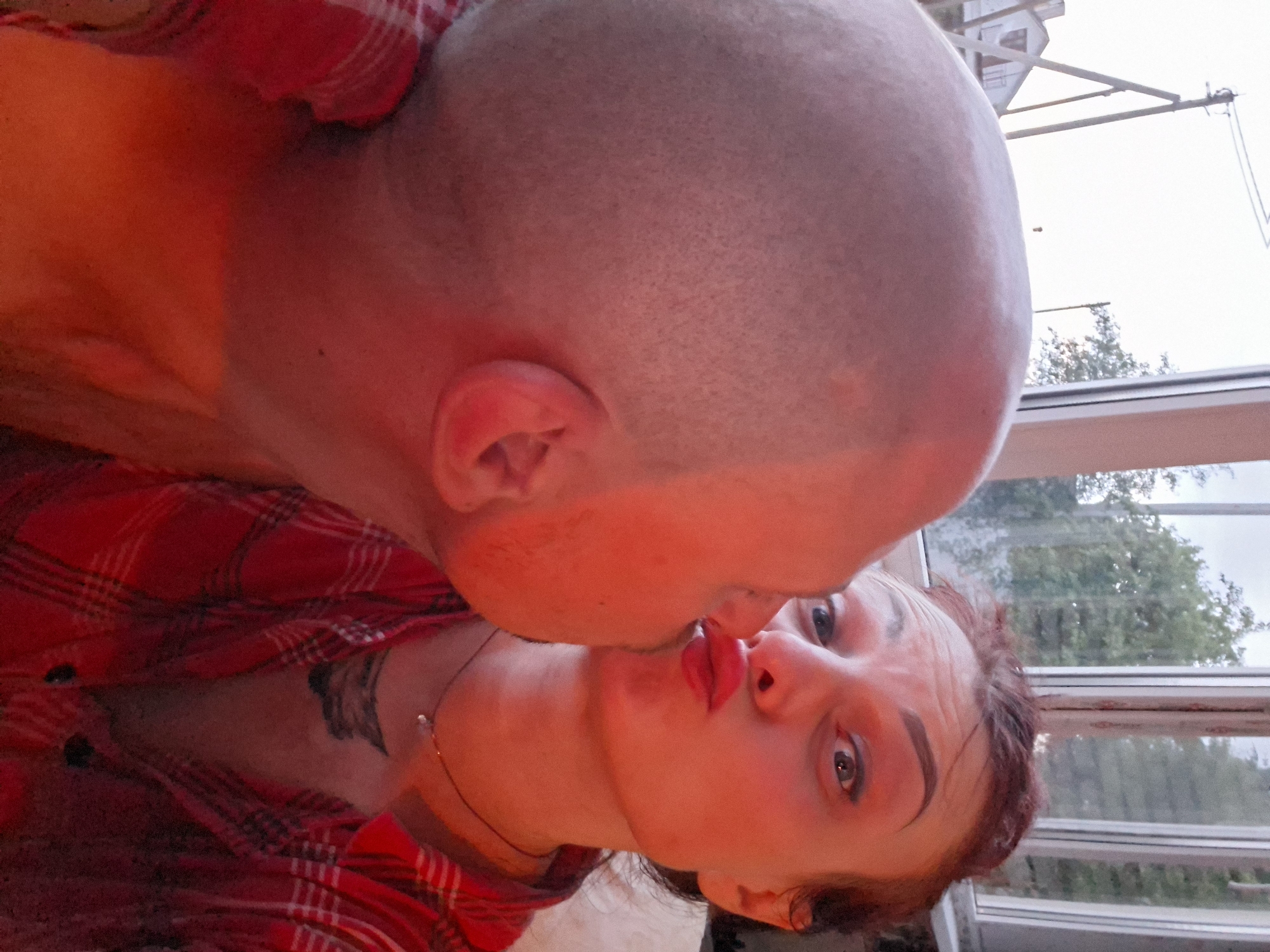 A close-up, over-the-shoulder selfie depicts two individuals, one in the foreground and the other in the background. The person in the foreground has a shaved head and is positioned to the left of the frame, with their head tilted towards the right. The person in the background, on the right, is a woman with reddish-brown hair, wearing a red and black plaid shirt. She is leaning in, puckering her lips as if to kiss the person in the foreground, whose head is the primary focus. Her eyes are wide open, and she appears to be looking directly at the camera. A thin necklace with a small pendant is visible around her neck, and a tattoo is partially visible on her upper chest. The lighting in the image is warm and reddish, casting a glow over both individuals. The background reveals a window with a view of trees and a building structure, suggesting an outdoor or semi-outdoor setting. The style of the window and the distant building hints at a location in a rural or suburban area. Given the metadata, the setting is likely in Забалоцкі сельскі Савет, Belarus.