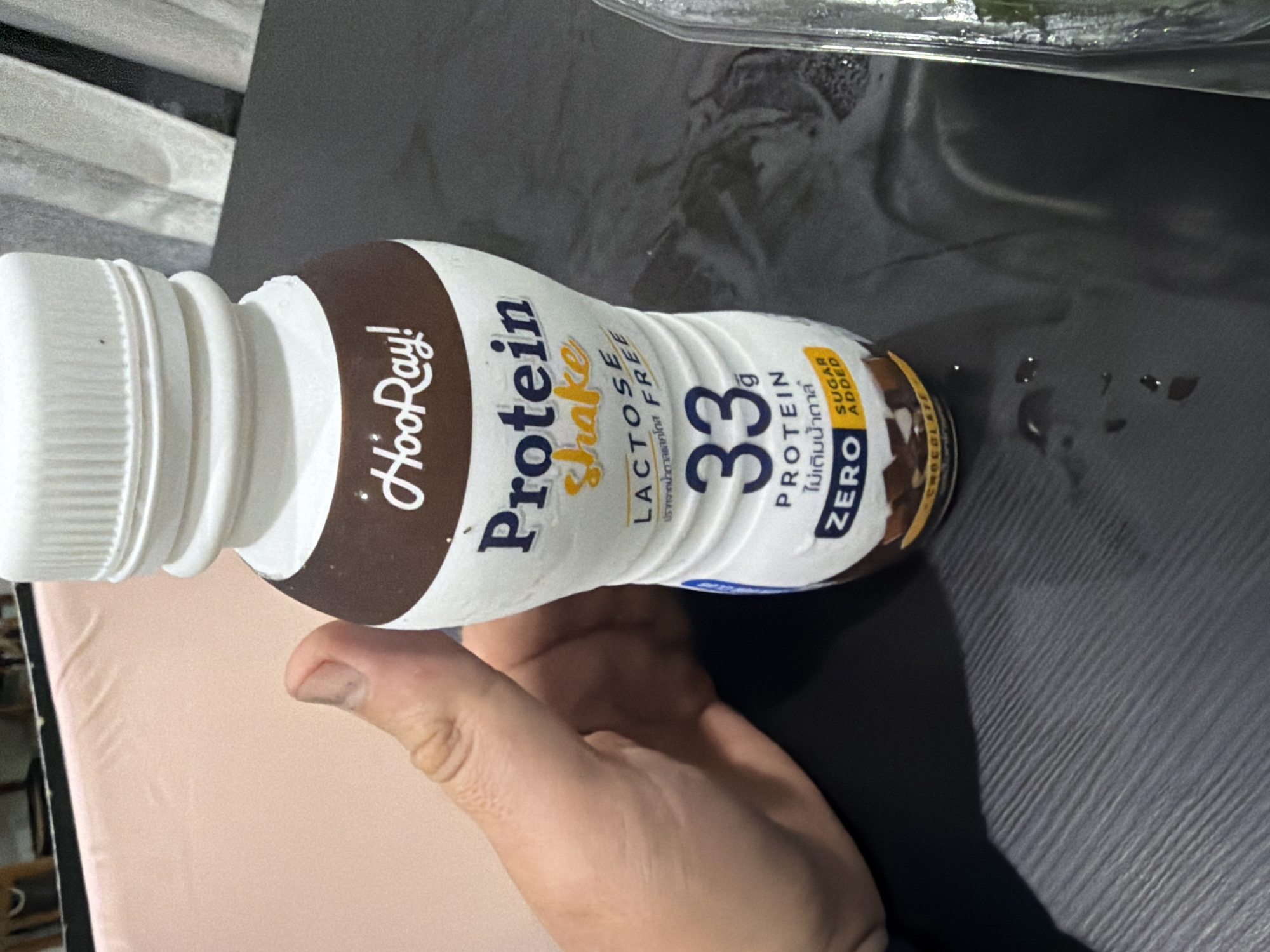 A hand is holding a bottle of HooRay! Protein Shake in Baan Mae Nam, Thailand. The bottle is white and brown, with "Protein Shake", "LACTOSE FREE", "33g PROTEIN", "ZERO SUGAR ADDED", and "CHOCOLATE" printed on it. The background is a dark, textured surface.