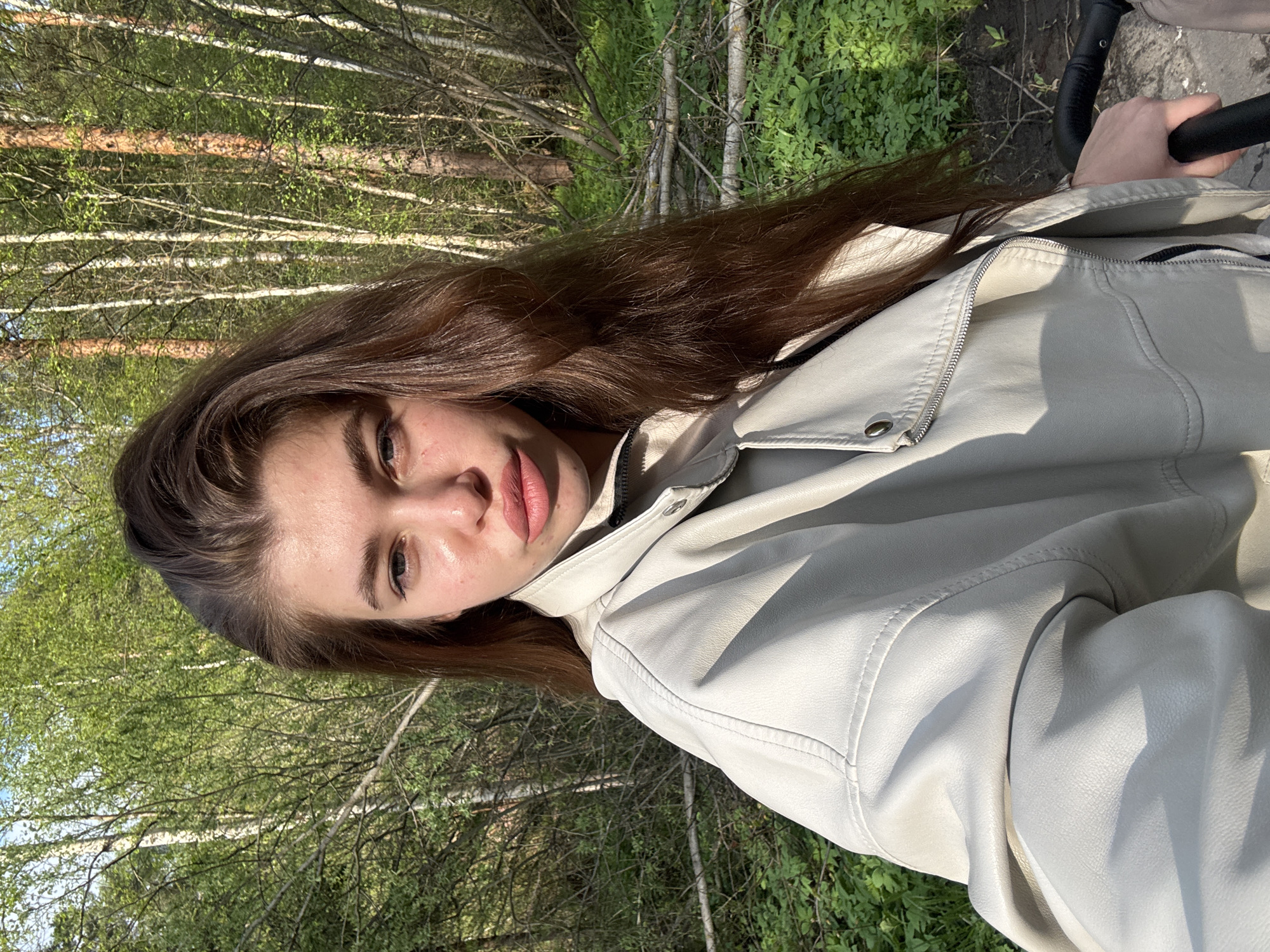 A woman in a light-colored jacket is photographed in a forest. The forest setting is suggested by the presence of trees with light-colored trunks and green foliage. The woman has long brown hair and is positioned at an angle in the frame. She appears to be holding a black handle, possibly from a stroller or similar object. The location is the city of Мещёры, Russia.