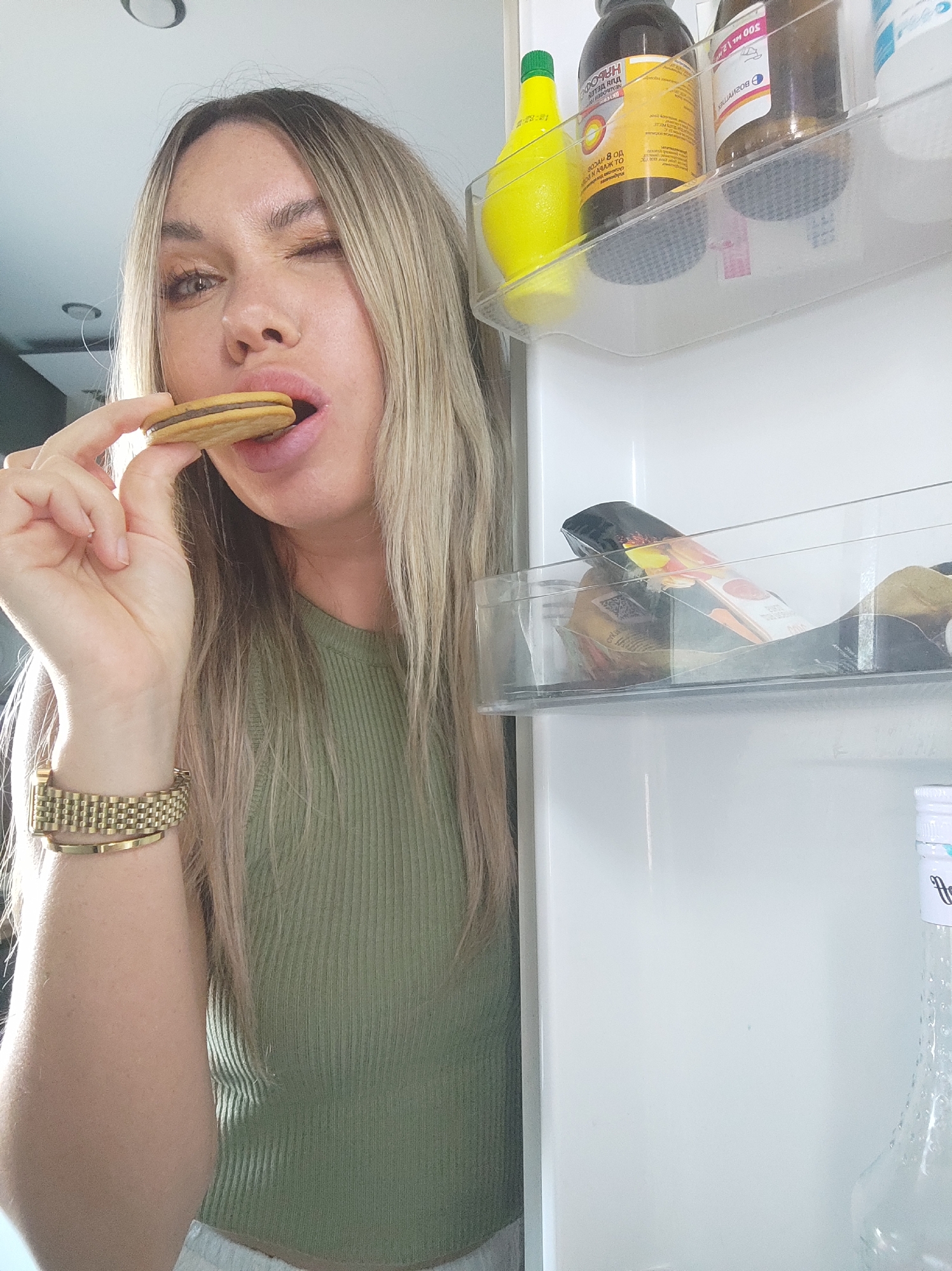 A woman is in front of an open refrigerator. She is fair-skinned, with blonde hair, and is wearing a green top and a gold watch. She is winking and holding a sandwich cookie to her mouth. Inside the refrigerator are bottles of liquid on the top shelf, and packaged food on the shelf below. The location is not discernible as Votkinsk, Russia.