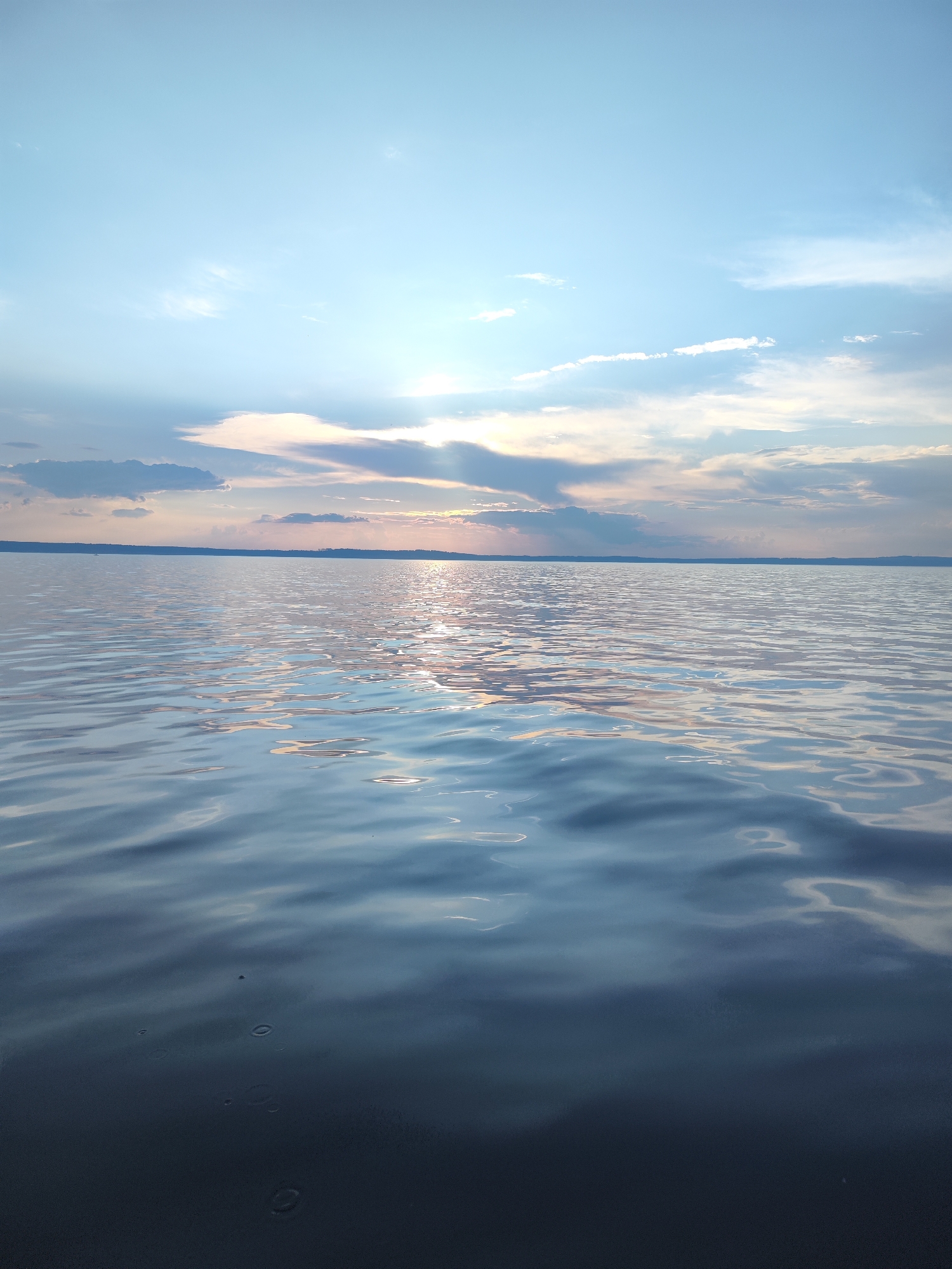 The image depicts a serene body of water, likely a lake or a large river, under a partly cloudy sky. The sky is a pale blue with scattered white and gray clouds, some of which are illuminated by the setting sun, casting rays of light that are visible through the clouds. The sun itself is not directly visible but its presence is indicated by the bright reflection on the water's surface. The water is calm with gentle ripples, reflecting the colors of the sky and clouds. The horizon line is visible, separating the sky from the water, and a faint, dark landmass can be seen in the distance. The foreground of the image shows the water in more detail, with the ripples creating patterns of light and shadow. Several small, circular disturbances are visible on the dark surface of the water in the lower part of the image, possibly from raindrops or small creatures. There are no people, boats, or structures visible in the image. The scene appears to be in a natural, undeveloped area, and the location in the city of Галево, Russia, suggests a possibly tranquil, outdoor setting near a body of water within or adjacent to the city.