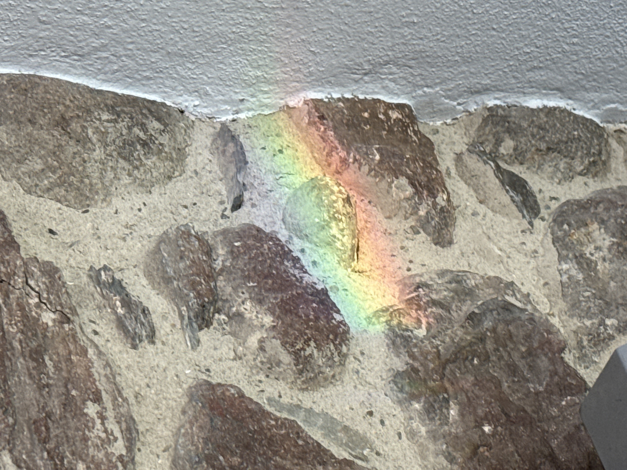 The image shows a close-up of a stone wall with a rainbow reflection on it. The wall appears to be made of irregularly shaped stones of various sizes, held together by mortar. The stones are primarily grey and brown in color. The rainbow is located in the center of the image, stretching across the stones, displaying the full spectrum of colors. The wall is situated under a white surface, possibly a ceiling or an overhang. The image likely depicts a detail in the city of Benasque, Spain, considering the request.