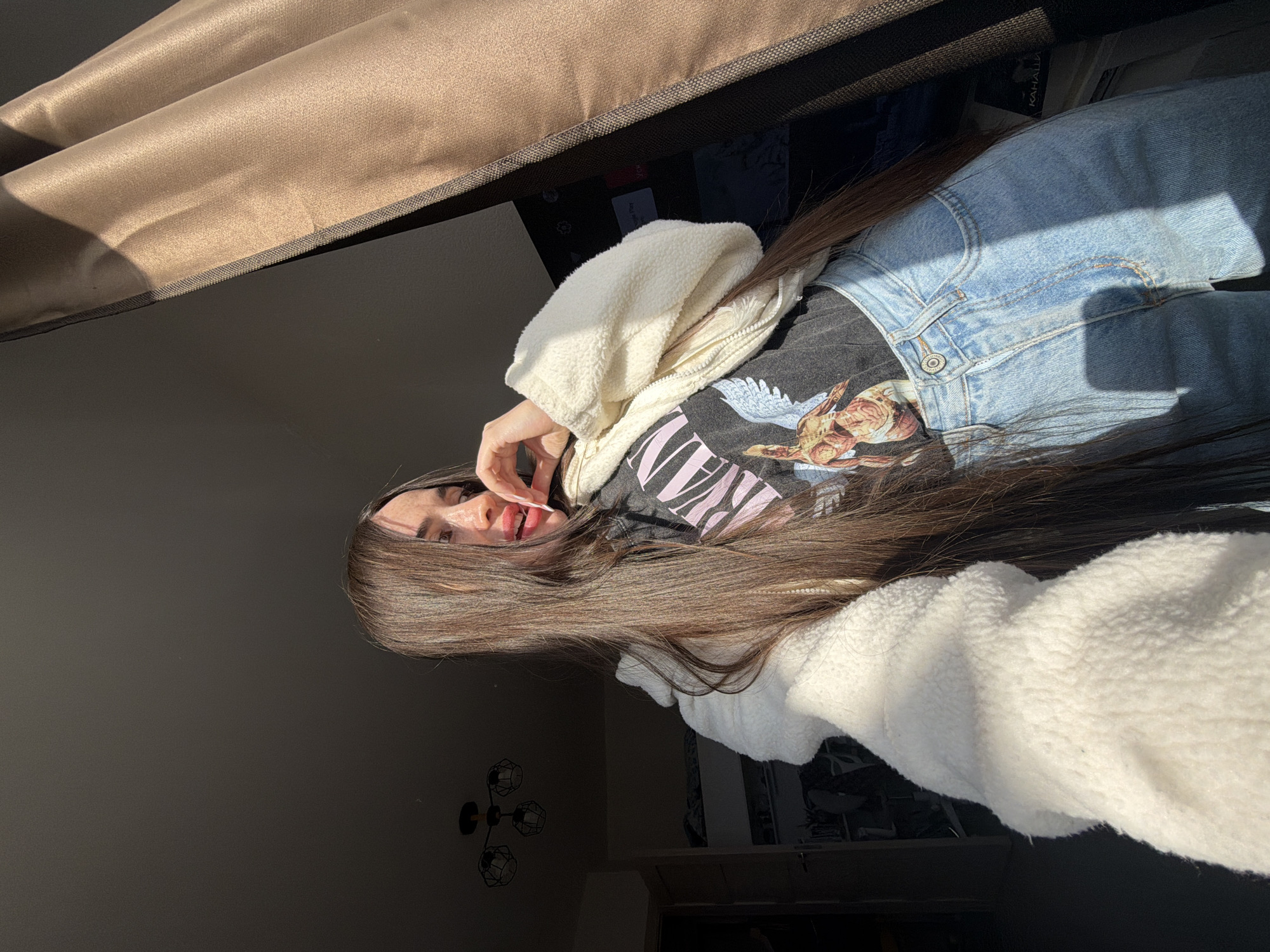 A female subject is depicted indoors in a selfie-style, eye-level photograph, located within an interior in Saint Petersburg, Russia. She is positioned slightly off-center to the left, with her upper torso and head angled to the left, while her gaze is directed generally forward. Her long, dark brown hair cascades over her shoulders and chest. She wears a black t-shirt with a partially visible graphic design, light-wash blue jeans, and an open cream-colored, fluffy zip-up jacket. Her left hand is raised to her face, with red-tipped fingers touching her lips and chin.
Strong natural sunlight enters from the right, creating harsh highlights on the subject and casting deep shadows in the background. The background on the left reveals a dimly lit room with a dark ceiling and a modern ceiling light fixture featuring three geometric wireframe shades. A dark doorway further back shows a glimpse of what appears to be a bunk bed structure. The background on the right is dominated by a heavy, brown curtain, partially illuminated by sunlight. Below the curtain and to the right of the subject, a large, dark flat-screen television or monitor is partially visible. An object with the Russian word "КАНАЛЫ" (Channels) is discernable on a surface in front of the television, confirming the Russian location.