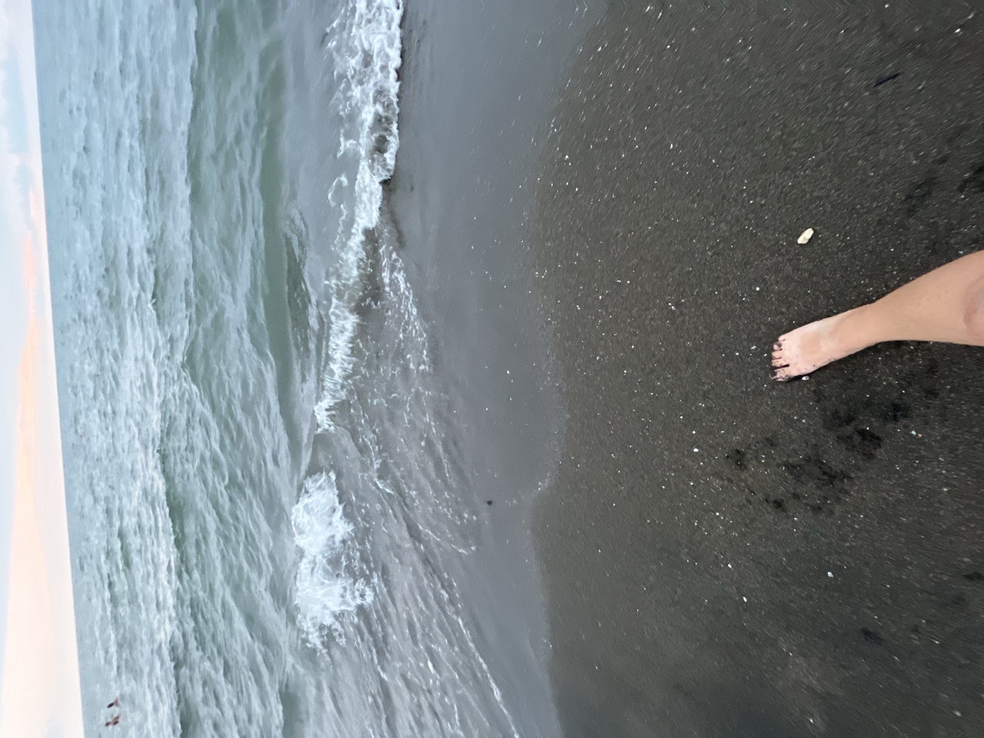 The image shows a person's bare foot on the dark sand of Ureki, Georgia's Black Sea coast. The foot is positioned in the lower right quadrant of the frame, with the toes pointed towards the left. The skin of the foot and lower leg is visible, appearing lightly tanned. The sand is dark and speckled with small white shells or pebbles. Gentle waves are breaking on the shore in the mid-ground, creating a foamy white edge. The water is a murky greenish-blue, with ripples on the surface. In the far distance, the sky is visible with a faint pink and orange hue, suggesting either sunrise or sunset. Two figures are faintly visible in the water in the upper left of the image, appearing to be swimming. The overall environment is a natural beach scene at Ureki, Georgia.