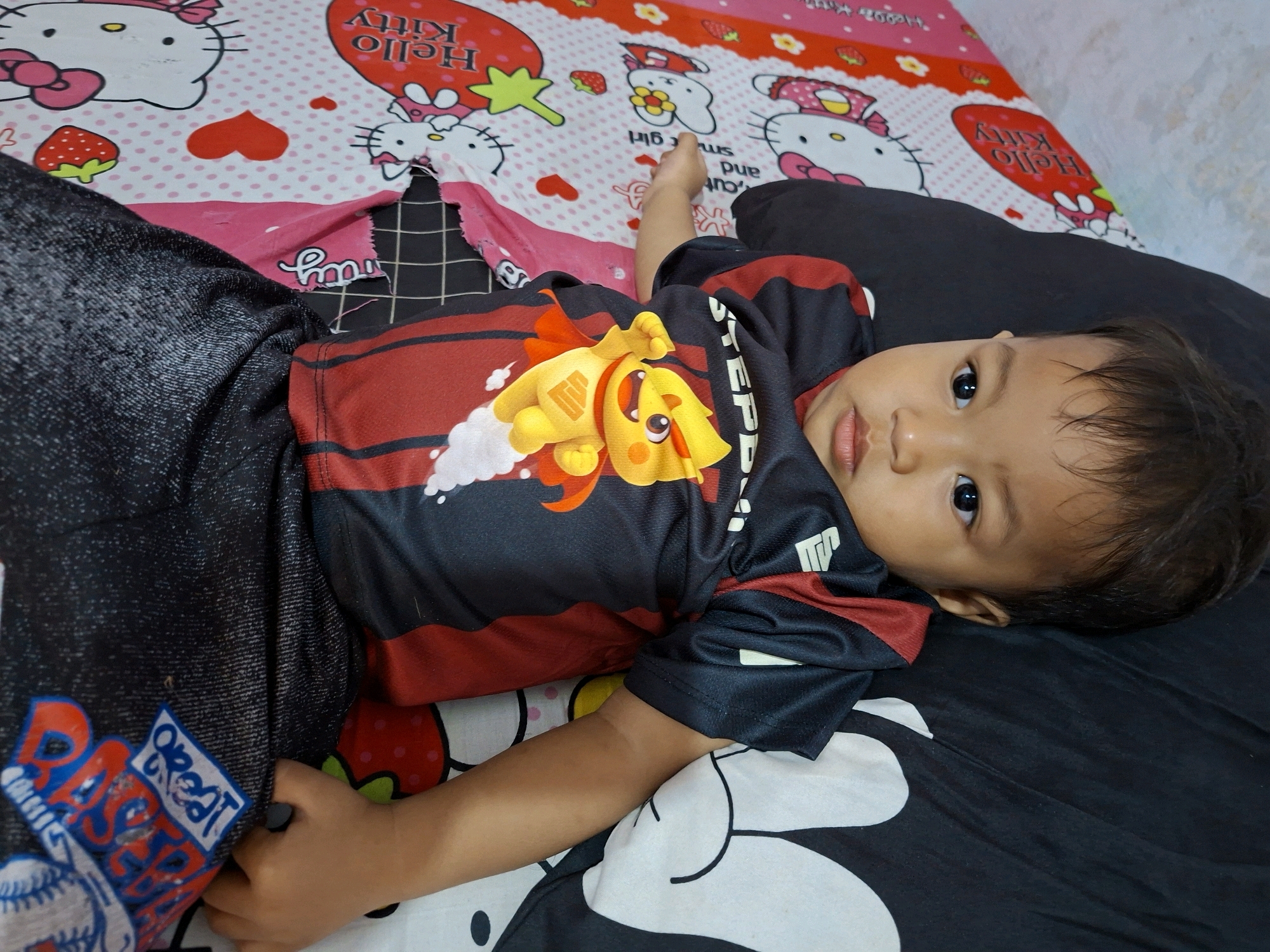A young child, likely a toddler, is lying on their back on a bed. The child is wearing a black and red t-shirt with a graphic print featuring a yellow cartoon character with a cape. The child's legs are covered by dark pants, possibly jeans.
The bedspread is decorated with a pattern of cartoon characters, specifically Hello Kitty, along with hearts and strawberries. A portion of the bedspread appears to be torn or bunched up, revealing a black and white checkered pattern underneath. The child is looking directly at the camera with a neutral expression.
The lighting suggests it is daytime, and the indoor setting is confirmed by the bed. The presence of the cartoon-themed bedspread indicates a home environment, possibly a child's bedroom. There is no visible text within the immediate focus of the child or their clothing, though the bedspread has some stylized lettering that is not clearly legible. The overall scene conveys a sense of a casual, relaxed moment.
