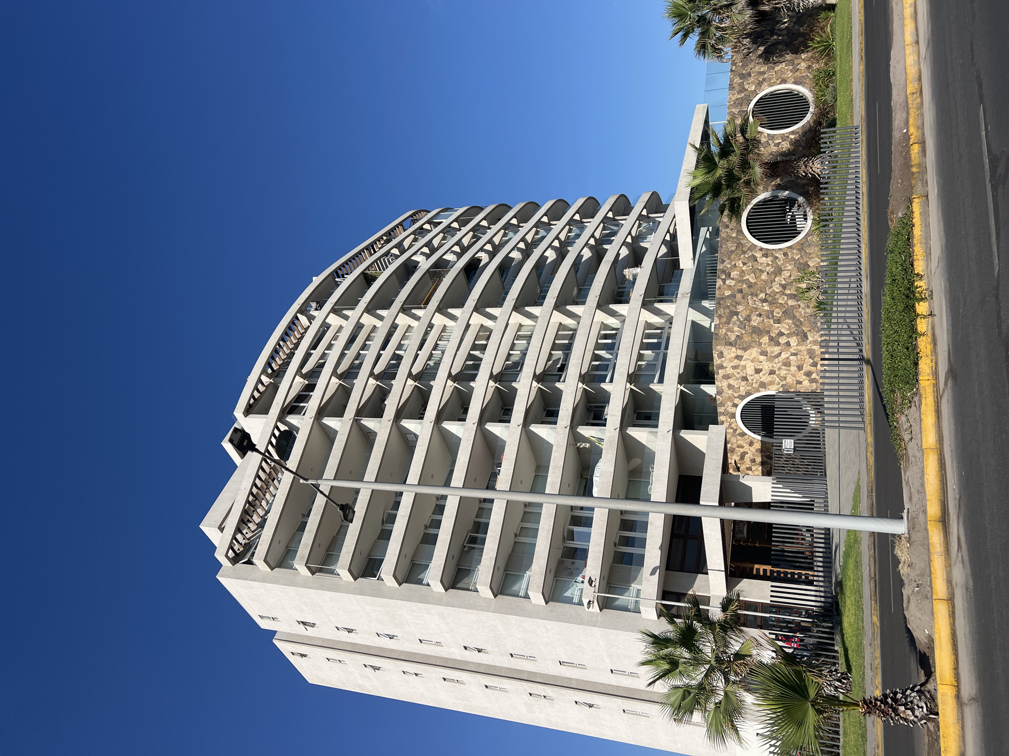 The image shows a tall, modern apartment building in Arica, Chile, angled against a clear blue sky. The building features a curved facade with repeating concrete arches supporting balconies. The lower portion of the building is a flat wall. To the right, a stone wall section with three circular ventilation ports is visible, bordered by a metal fence and a sidewalk with a yellow curb. A palm tree stands in front of the building. A street lamp is positioned in front of the building. A narrow asphalt road runs alongside the building, bordering the sidewalk. People are visible on the balconies and in the shadow under the front of the building.