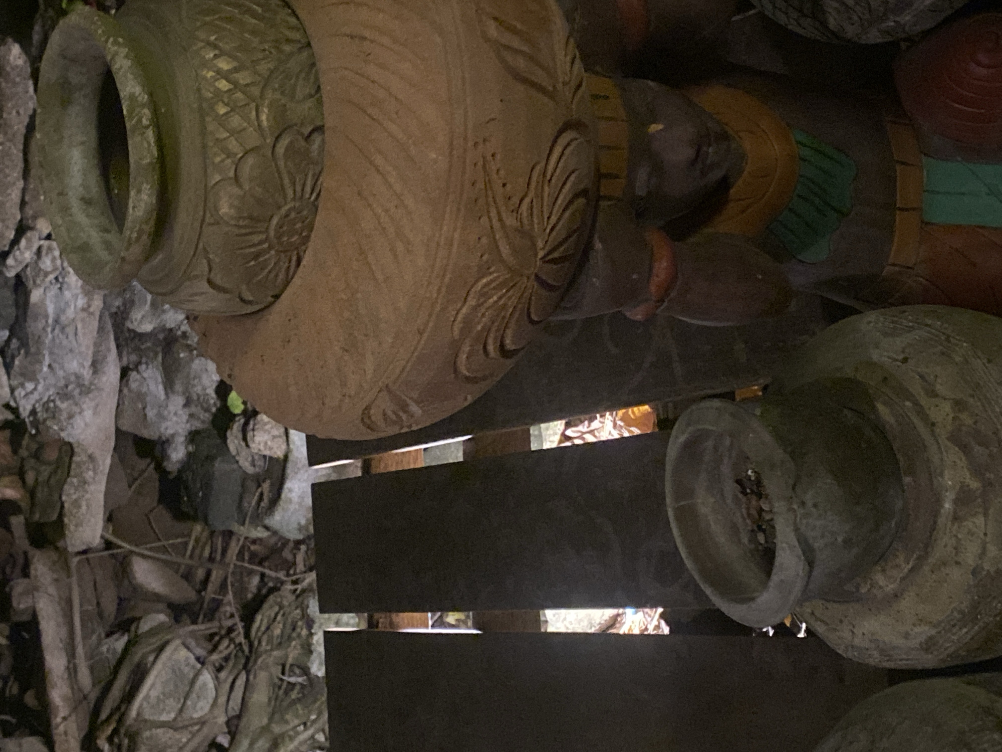 The scene is outdoors, likely in a garden or outdoor display area in Baan Don Sai, Thailand. The focal point is a collection of large, ornate terracotta pots. One prominent pot is light brown with carved floral patterns around the top. Another pot, grey and weathered, shows its interior filled with pebbles or dirt. These pots rest on dark wooden slats or a platform, with visible space between the slats revealing leaf litter and debris underneath. The background shows rocks, roots, and other ground covering materials. A portion of a decorative statue is also visible, featuring painted sections in yellow, green, and red. The lighting appears dim, perhaps during the evening or under heavy shade.
