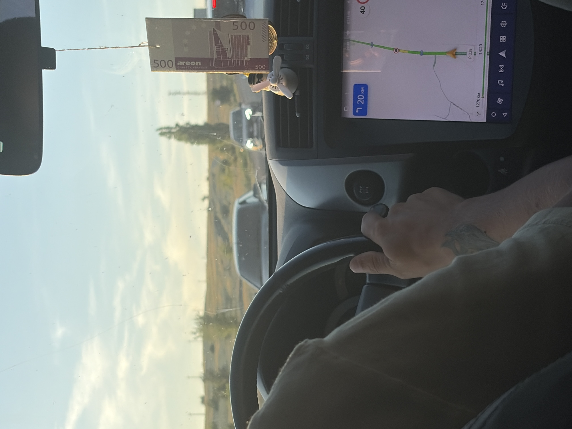 The image shows the interior of a car driving on a road, likely in Belogorki, Russia, given the context provided. A person's hands are visible on the steering wheel, indicating they are driving. The dashboard features a large touchscreen navigation system displaying a map with a route and distance. A decorative air freshener hangs from the rearview mirror. Outside the car, the sky is clear with some clouds, and there are other vehicles on the road ahead, suggesting moderate traffic. The surrounding landscape appears to be rural or semi-rural, with hills and sparse vegetation visible.