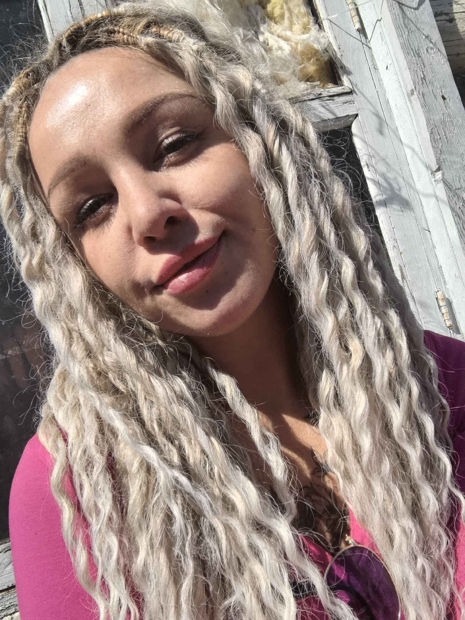 A selfie of a person with light-colored, braided hair wearing a pink top. The background includes a white, weathered wooden structure, possibly a building or fence, with exposed insulation visible through a hole. There is no clear indication of the location being Noviy, Russia.