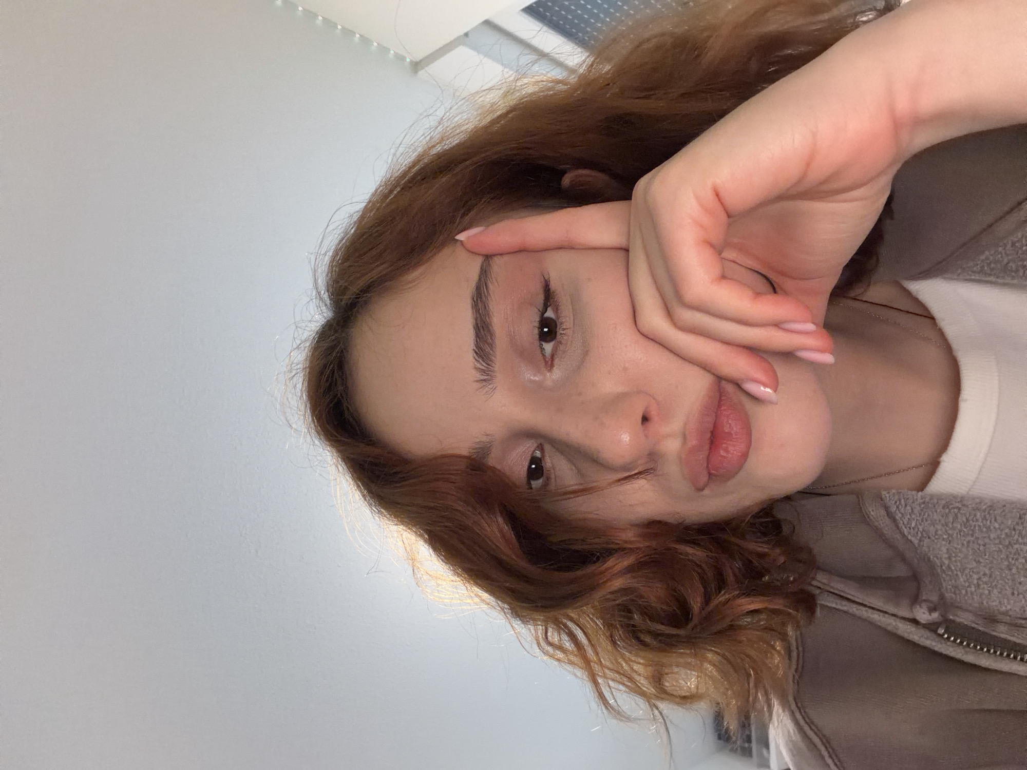 This is a close-up selfie of a young woman with reddish-brown wavy hair. She is looking towards the camera with her right index finger touching her forehead and her left hand resting on her cheek. Her lips are slightly parted, and she has light makeup on, including blush and eyeliner. She is wearing a gray zip-up hoodie over a white t-shirt and a thin silver necklace. The background is out of focus, showing a light-colored wall and a hint of a window with blinds. There is no indication of the location being in Strasbourg, France.