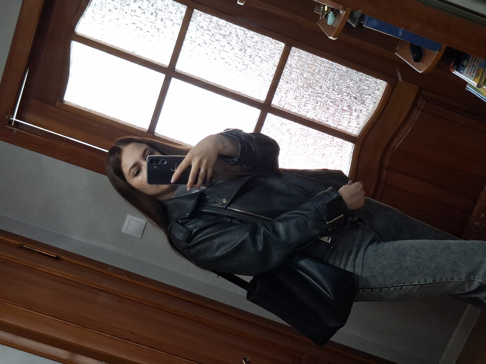 A young woman is taking a selfie in front of a mirror. She is wearing a black leather jacket and grey jeans. The background includes a wooden-framed window with patterned glass and a wooden surface that is likely part of a cabinet or wall paneling. The presence of books on a shelf suggests the location could be a home or study. There is no evidence to confirm that the location is Chaikovsky, Russia.