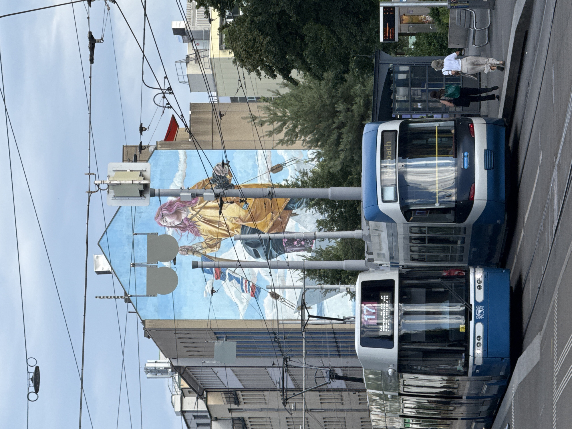 The image depicts a street scene in Zurich, Switzerland, with a large mural on the side of a building. Two blue and white trams are parked side-by-side on tram tracks. The tram in the foreground displays "17" on its digital display and the word "Seebach" on another display. The tram on the right is partially visible. Two women are walking on the sidewalk next to the tram on the right, with one carrying a green backpack and the other a tote bag. Overhead, a complex network of wires for the trams crisscrosses the sky. The mural on the building features a stylized depiction of a person with pink hair, wearing a yellow coat, holding objects, against a backdrop of clouds and flags. The building itself is light blue. Other buildings with windows and architectural details are visible behind the mural. Trees are present on the right side of the image. A bus stop with a shelter and benches is situated near the tram tracks. A digital information board is also visible at the bus stop.