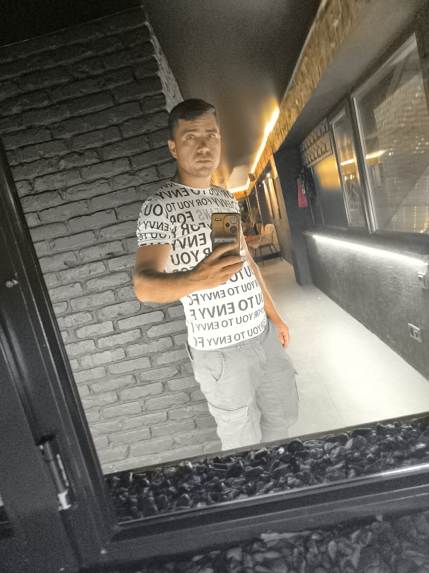 A man stands in front of a brick wall, taking a selfie in a mirror. He is wearing a white t-shirt with black text and grey cargo shorts. The mirror reflects his image and the environment behind him, which appears to be an indoor space with lighting and a hallway. The brick wall in front of him is dark grey. The floor in front of the mirror is a mosaic of dark stones. The image is taken in the city of Vulcănești, Moldova.