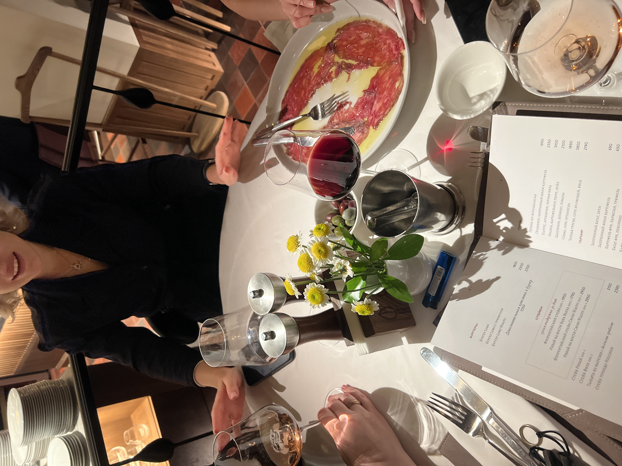 The image shows a table setting in what appears to be a restaurant. There are at least two people dining, indicated by their hands and partial view of a face. The table has a white tablecloth, on which are placed: a large oval plate of thinly sliced meat (possibly carpaccio) with oil; a glass of red wine; a glass of pink wine; a small white bowl; cutlery (forks and knives); a menu; a small vase with white and yellow flowers; and salt and pepper grinders. The menu is in Russian, suggesting a location in Russia, and references “Moscow” in one of the entries, indicating the city. A stack of white plates can be seen to the left. The lighting is subdued, and the overall atmosphere suggests a fine dining establishment.