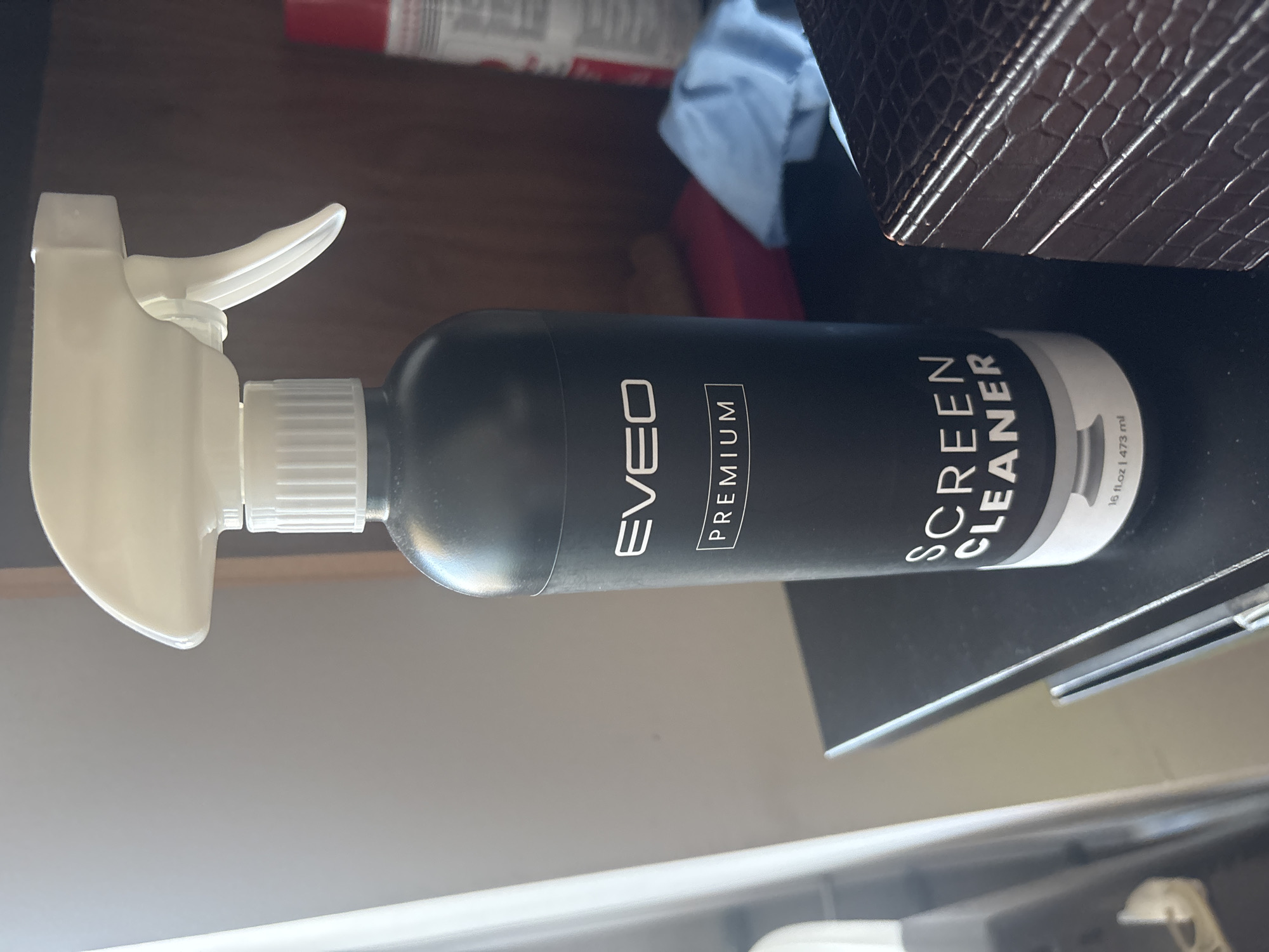 A black spray bottle of "EVELO PREMIUM SCREEN CLEANER" stands on a dark, possibly wooden, surface. The bottle has a white spray nozzle and cap. The label on the bottle is white text on a black background and reads "EVELO PREMIUM SCREEN CLEANER" with "16 fl.oz | 473 ml" at the bottom. In the background, a red and white can is visible on the right, and a blue microfiber cloth is folded to the left of the can. The scene is indoors, in a location that cannot be determined from the image.