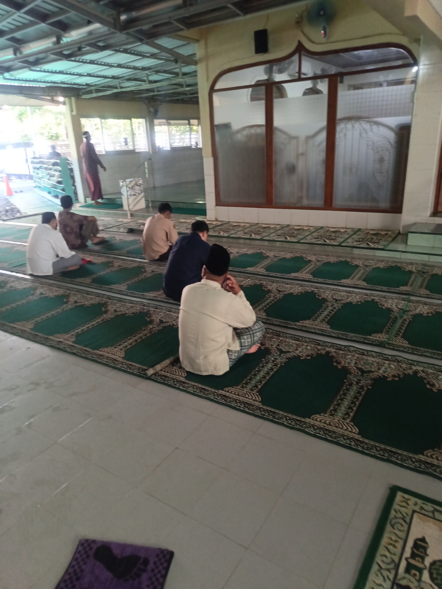 The image depicts the interior of a mosque in Desa Gunungsimping, Indonesia. Several men are seated on green prayer mats laid out on the floor. The mats have a patterned border. In the background, there is a large window with decorative glass panels and wooden framing. Beyond the window, the ceiling structure of the mosque is visible, supported by metal beams. To the left, there is a person standing in what appears to be a maroon robe, and further left, a row of shoe racks. The floor is tiled. A small purple mat with a heart design is visible in the foreground on the tiled floor.