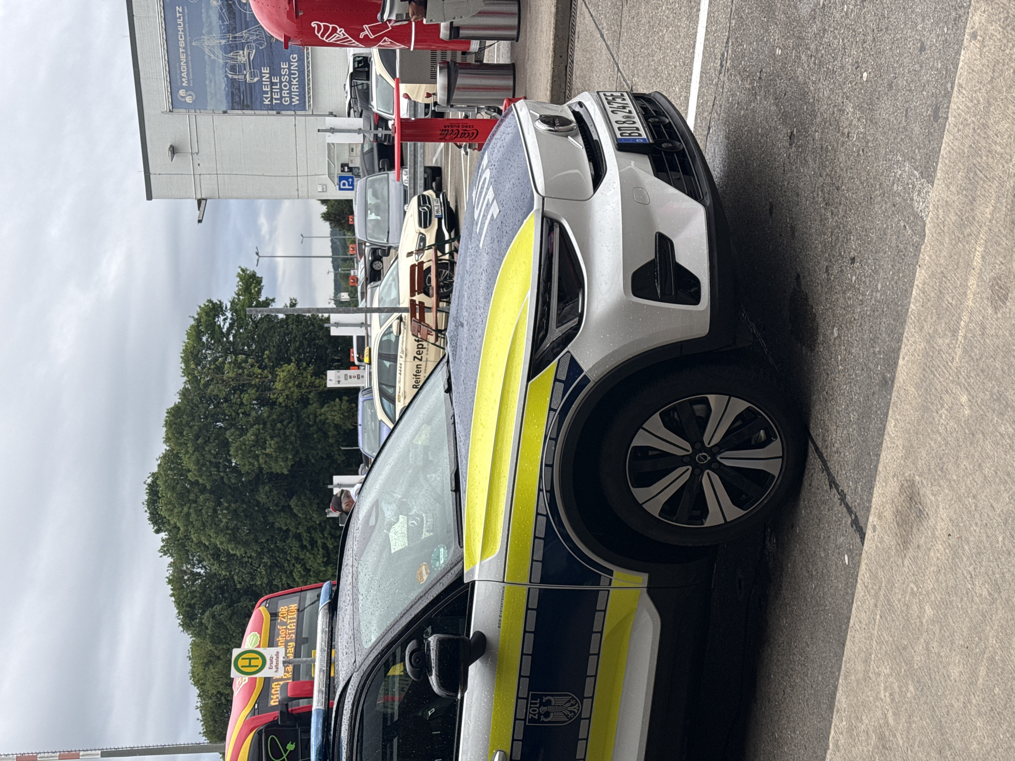 This vertical shot captures an outdoor scene in Künersberg, Germany, likely a parking area or a roadside stop on an overcast day, indicated by the gray sky and raindrops visible on vehicles. The central subject is a white, blue, and bright yellow German Zoll (customs) vehicle, parked with its front facing right. Its license plate, "BD8 2475E", is clearly visible on the front bumper. The car shows visible water droplets from recent rain or drizzle.
To the left of the Zoll vehicle, a red bus with a prominent yellow "H" (Haltestelle - bus stop) sign is visible. Its digital display reads "Ersatzhaltestelle" (replacement bus stop) and "Inhof ZOB Railway STATION," suggesting it might be serving a temporary route or a central bus station near a railway.
In the background, behind the Zoll vehicle and bus, several other cars are parked. A large white building features an advertisement for "MAGNETSCHULTZ" with the slogan "KLEINE TEILE GROSSE WIRKUNG" (Small Parts, Big Impact). Below this, a red Coca-Cola dispenser is visible. Further back, green trees line the horizon under the cloudy sky. Another sign, partially visible in the background, mentions "Reifen Zepf" (Zepf Tires). A blue "P" parking sign is also present on a pole. No people are distinctly visible, and no specific activities beyond parking and waiting at a bus stop are depicted. The overall mood is calm and mundane.