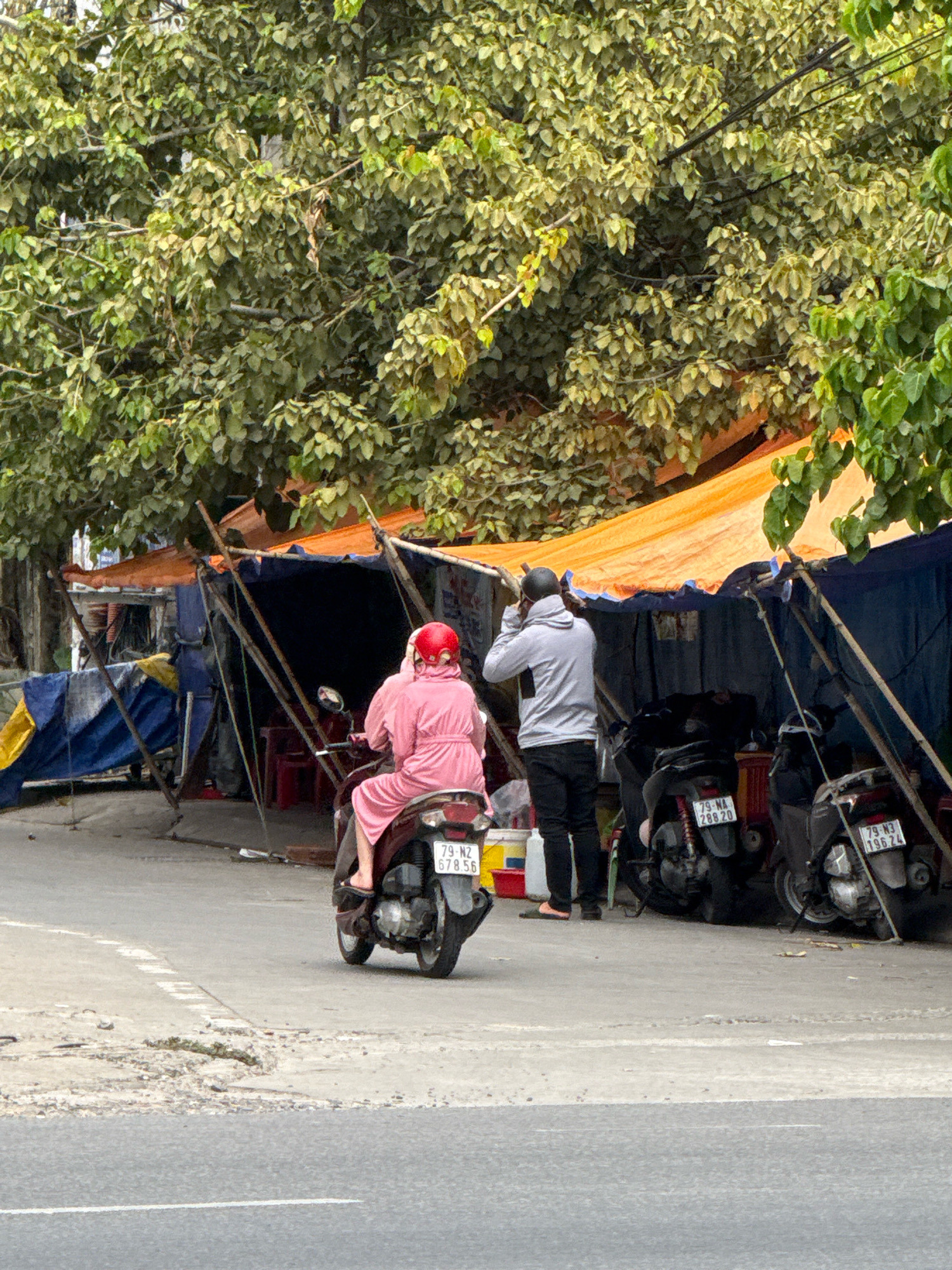 The image depicts a street scene in Nha Trang, Vietnam, with a focus on temporary market stalls set up along the roadside. In the foreground, a person wearing a pink robe and a red helmet is riding a motorcycle. Behind them, another individual in a grey hooded sweatshirt and black pants stands looking at their phone, positioned near a makeshift shelter constructed with blue and orange tarpaulins supported by wooden poles. Several motorcycles are parked in front of the stalls. The environment is characterized by lush green trees providing shade overhead and a weathered concrete road surface. The stalls appear to be selling goods, though the specific items are not clearly discernible. The overall atmosphere suggests a bustling, outdoor marketplace.