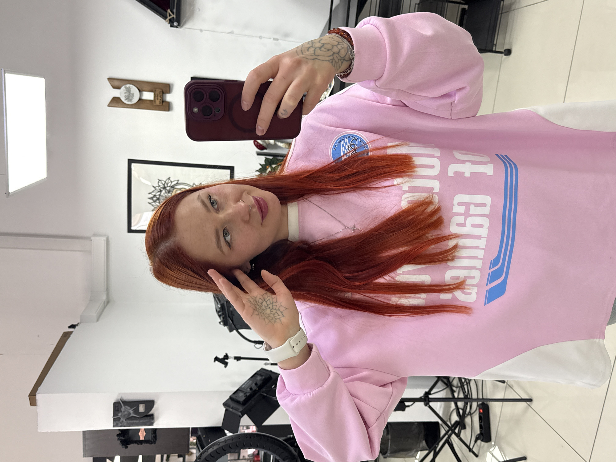 A young woman with long, vibrant red hair takes a selfie in what appears to be an indoor studio or professional space in Rostov-on-Don, Russia. She is holding a dark red smartphone in her right hand, positioned above her head, and looking directly at the camera with a subtle smile. Her left hand is gently touching her hair near her face.
She is wearing a light pink and white two-tone sweatshirt. The pink section features mirrored text in blue and white, which reads "CULTURE" or a similar word, with two blue horizontal stripes beneath. There's also a circular patch on her upper left chest with some indistinct white text and a blue checkered pattern, possibly saying "WIDE" and "MIDE" or "CRD." She has a silver cross pendant necklace, a nose piercing, and a white smartwatch on her left wrist. Notable tattoos are visible on her hands: a detailed mandala-like design on the back of her left hand, a smaller symbol on a finger, and another intricate pattern on her right wrist/forearm. A red beaded bracelet is also on her right wrist.
The background reveals a brightly lit interior with white walls. Visible objects include a framed artwork with a black and white floral or mandala design, a wooden wall decoration with a white circular emblem, and various pieces of studio equipment such as light stands, a ring light, a softbox (reflected brightly on the left), and a power strip on the floor. The setting suggests an environment possibly used for photography, video, or tattooing.