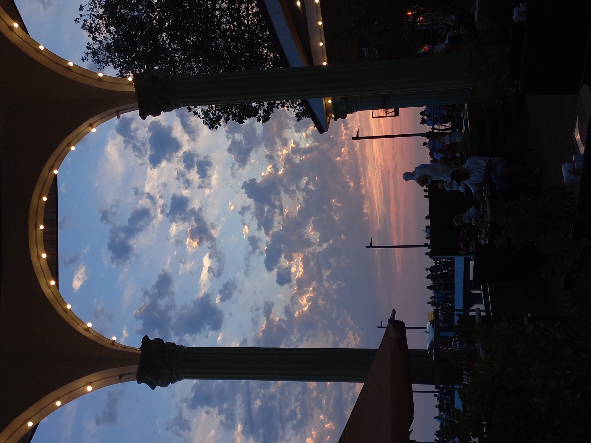 The image captures a serene dusk scene at the Sochi Arboretum, Russia. The sky is painted with hues of orange and pink as the sun sets, with scattered clouds catching the last rays of light. In the foreground, two large, ornate columns frame the view, adorned with string lights that add a warm glow. Beneath the arches, a statue of a woman holding a pot stands prominently, surrounded by smaller decorative figures. A crowd of people is visible in the background, some standing near a railing overlooking what appears to be a body of water, while others are seated at tables, enjoying the evening. The overall atmosphere suggests a peaceful and picturesque setting, with lush greenery and architectural elements contributing to the charm of the arboretum.