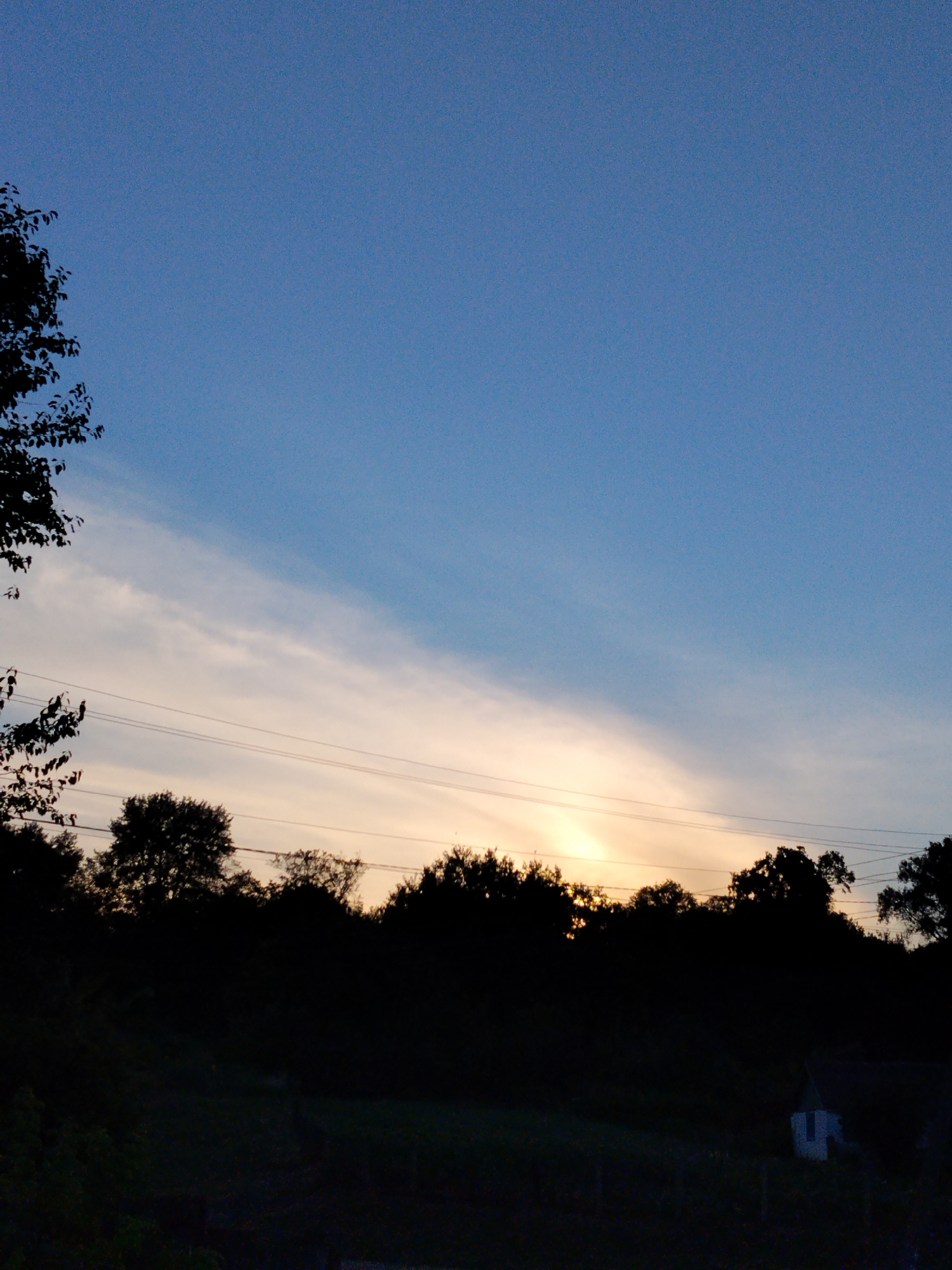 A silhouette of trees and a small white building against a twilight sky is visible. The sky is a gradient of deep blue at the top to lighter blue and yellow at the horizon. A thin band of clouds stretches across the sky. Power lines are strung between poles. The image was taken in Ustyvytsia, Ukraine.