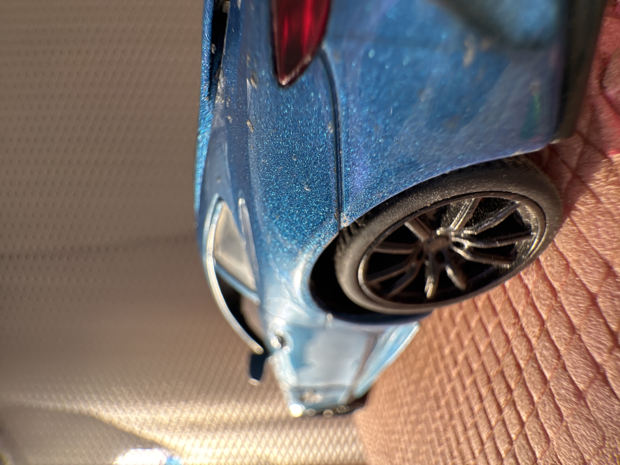 The image shows a close-up, angled view of a blue toy car, possibly a model car, with visible dust and debris on its surface. The rear of the car is in focus, showing the taillight and one of the wheels. The background is a textured, light-colored surface on the left, while the car is resting on a pinkish, fabric-like material on the right. The car is being held by a human hand. The exact location and the city of Novo-Nikol'skoe, Russia, are not discernible from the image.