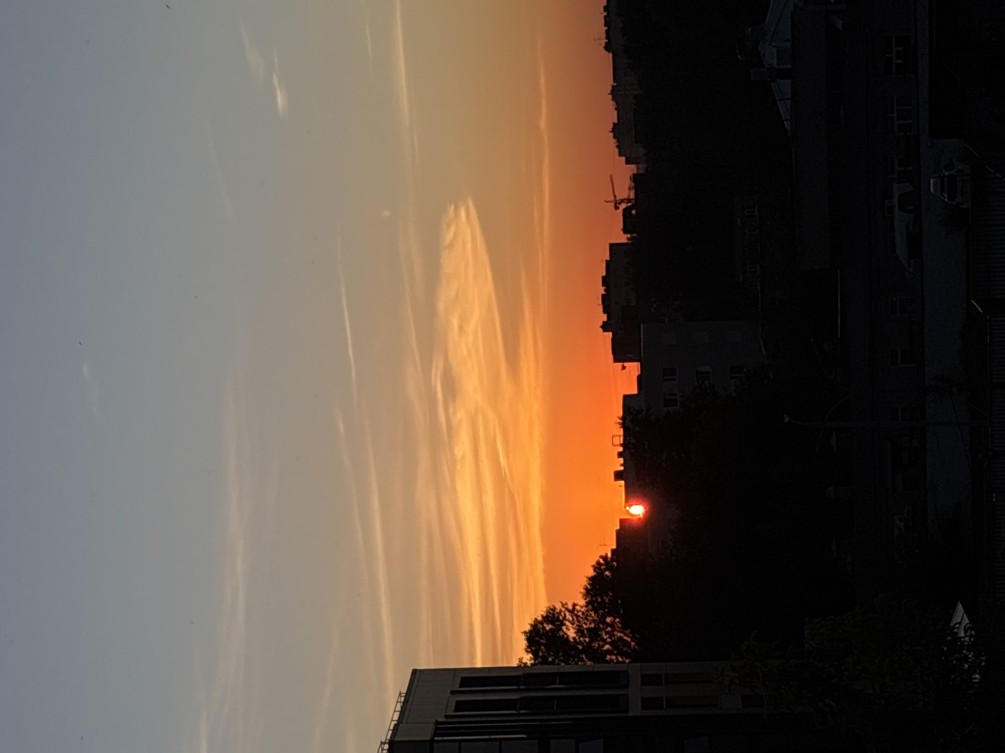 The image captures a sunset over a city skyline, likely Perm, Russia, based on the style of architecture. A bright orange sun is partially obscured by the silhouettes of trees and buildings. The sky transitions from a deep blue at the top to vibrant oranges and yellows near the horizon, with wispy clouds adding texture. In the foreground, the dark outlines of buildings and trees dominate, with a modern building visible on the left. The scene conveys a sense of urban twilight.