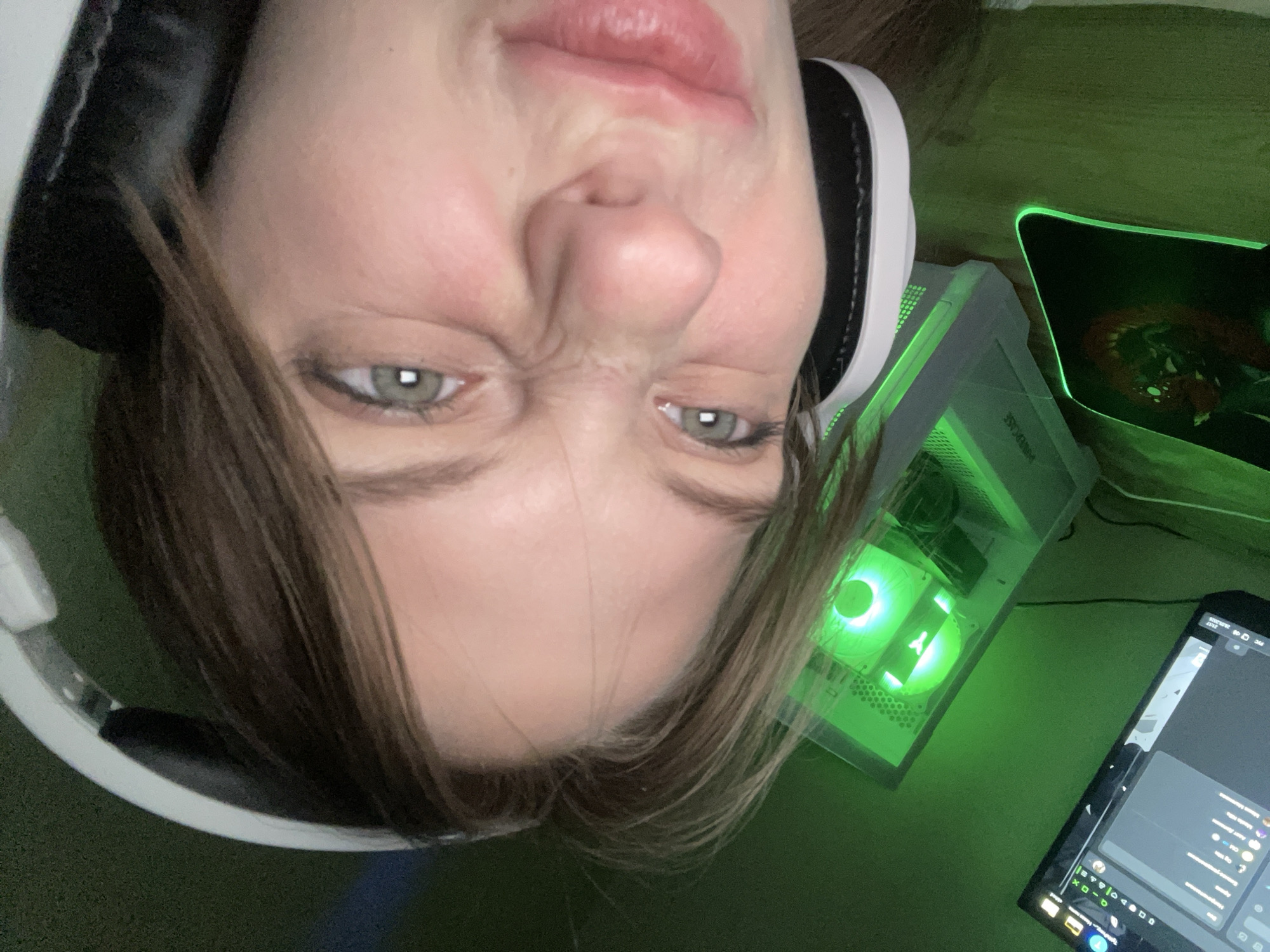 A person with light brown hair and green eyes is positioned in the upper left of the frame, wearing white headphones. The person is making a scrunched face. To the right of the person's head is a white computer tower with internal green LED lighting, a black mouse pad, and a black tablet displaying some software. The background is a solid green color. The image is oriented upside down. It is not possible to determine the location is Ufa, Russia.