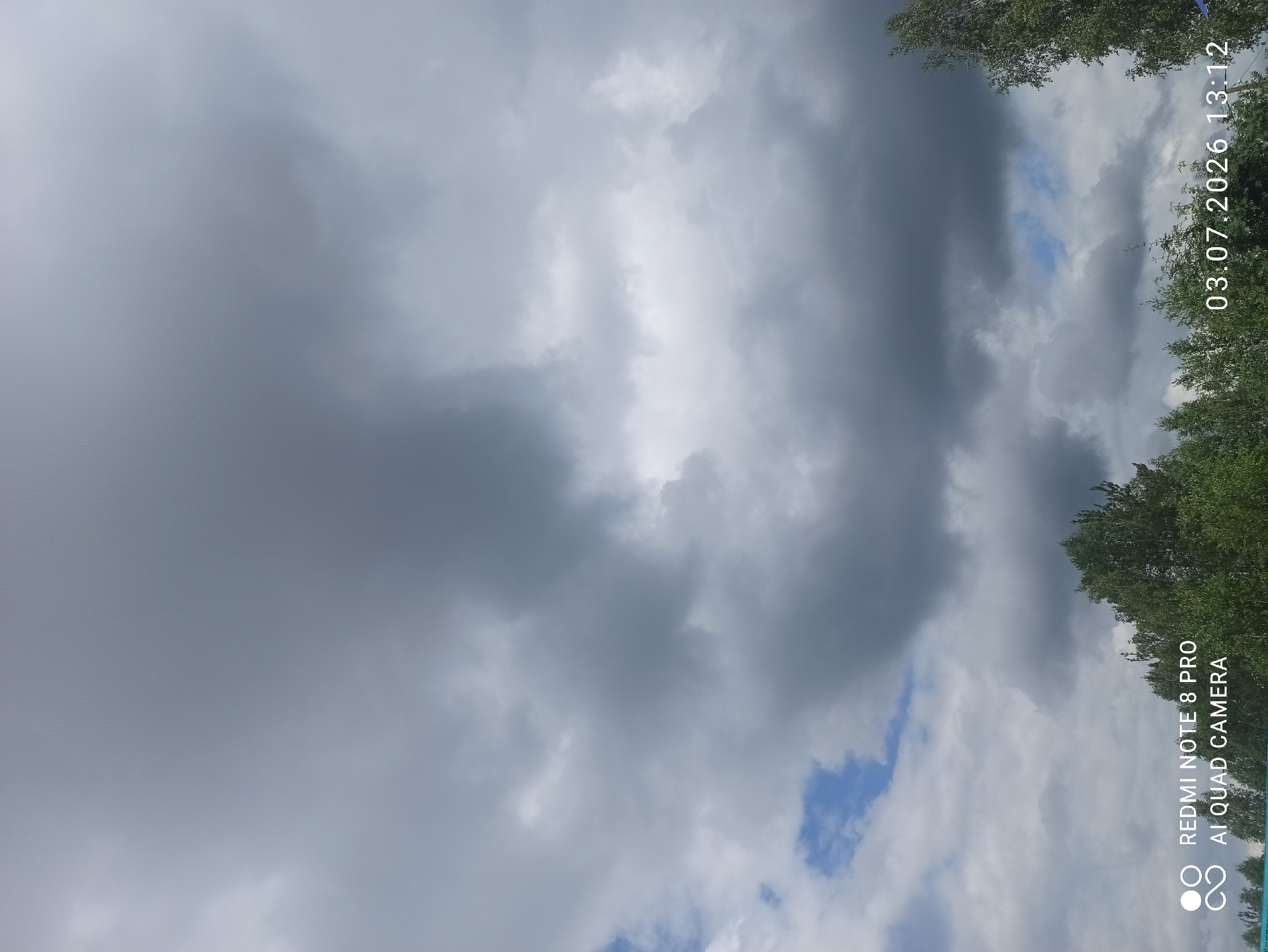 The image shows a cloudy sky over a treeline. The sky is filled with dark gray clouds, with some patches of blue sky visible. The treeline consists of green deciduous trees. In the bottom left corner, there is a watermark that reads "REDMI NOTE 8 PRO AI QUAD CAMERA." In the bottom right corner, a timestamp is visible: "03.07.2026 13:12." The image was likely taken in Sovetskiy, Russia, given the timestamp format which includes a year in the future, suggesting a potential placeholder or an error in the metadata.