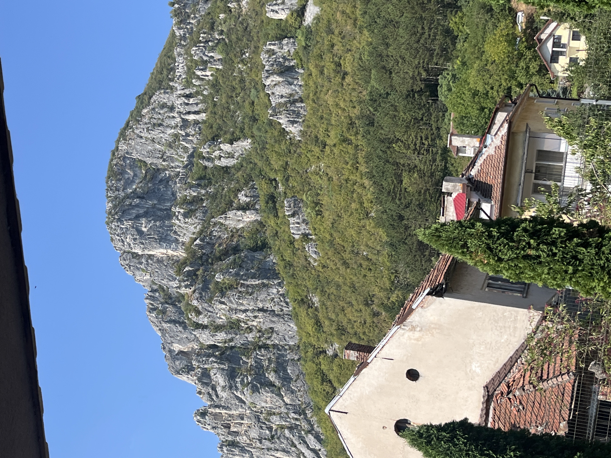 A view of a rocky mountain peak covered in vegetation and a few houses in the foreground, under a clear blue sky. The mountain is characterized by its rugged, gray rock formations and dense green foliage. The houses appear to be old, with terracotta tiled roofs and weathered facades. The scene likely depicts a location in or near Vratsa, Bulgaria, given the characteristic mountainous landscape. There are no people visible in the image.