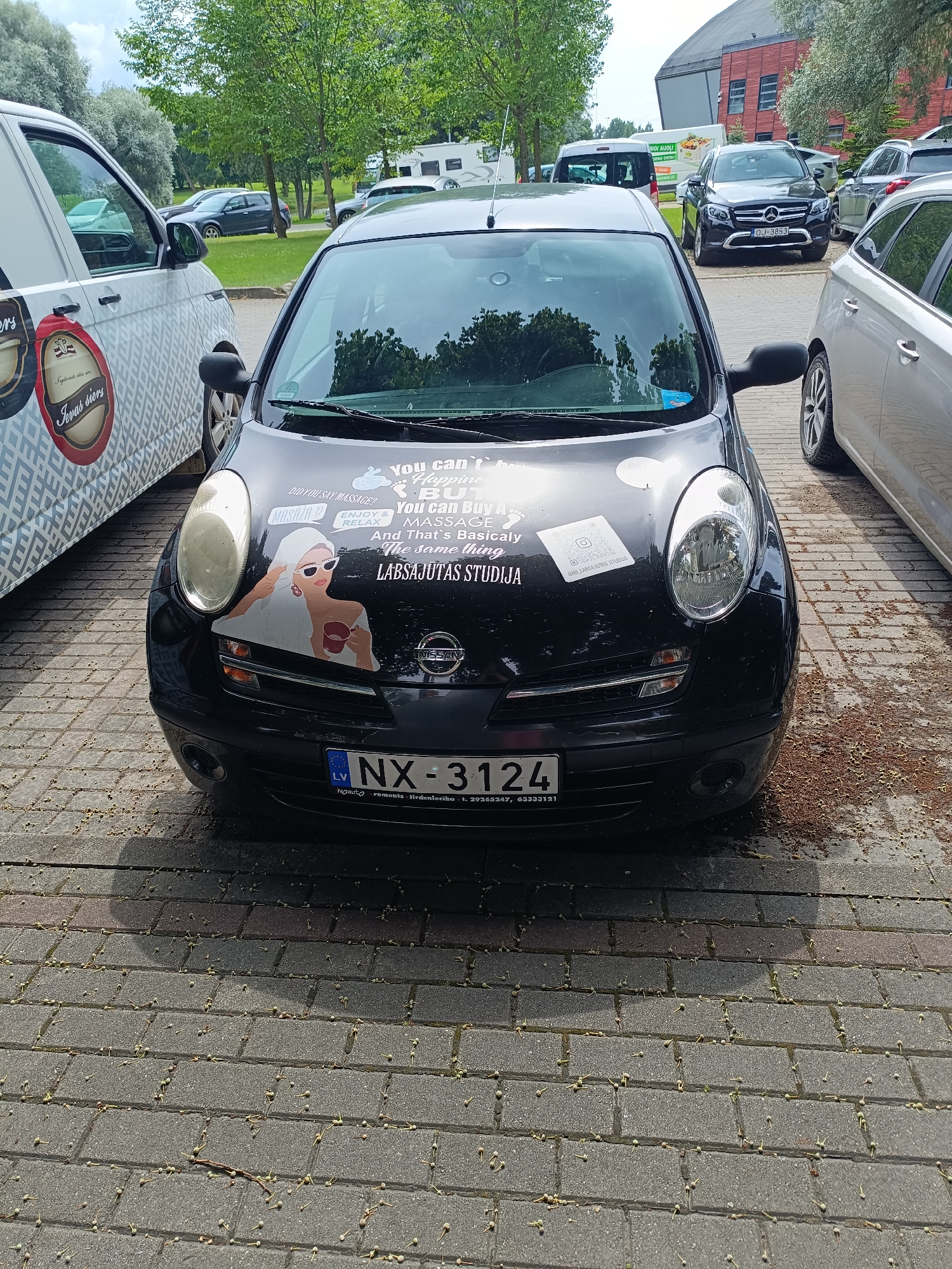 A black Nissan Micra is parked on a brick patio in Kuldīga, Latvia. The car has several stickers on its hood, including one of a woman holding a mug and another with a QR code. The license plate reads "NX-3124." To the left of the Nissan is a white van with a patterned design and logos. In the background, several other cars are parked, and trees and buildings are visible. The ground is covered in fallen leaves and small seed pods.