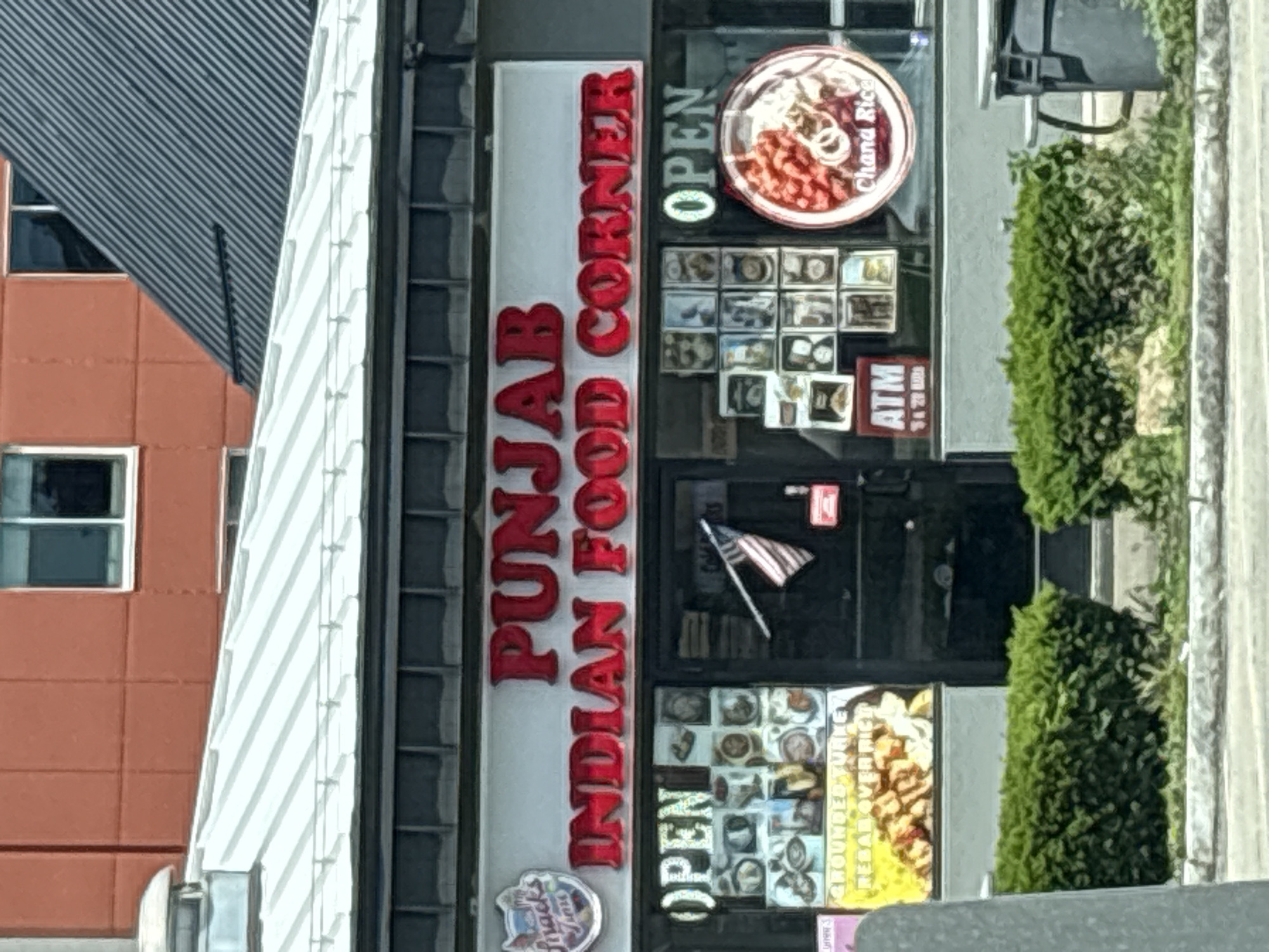 A storefront for "Punjab Indian Food Corner" is captured on a sunny day. The building features a metallic roof and a dark facade above the display windows. The restaurant's name is prominently displayed in large, red, three-dimensional letters. Below the name, the windows showcase various images of food, with "OPEN" signs visible on both the left and right sides. On the left, there is a display that includes the text "GROUNDED TURKEY REBAR OVER RICE". An American flag is displayed on a pole attached to the main entrance door. To the right of the door, a circular sign advertises "Chana Rice". An ATM is also visible near the entrance. In front of the building, well-maintained green bushes line the sidewalk. The environment suggests a commercial street in the city of Niagara Falls, United States of America.