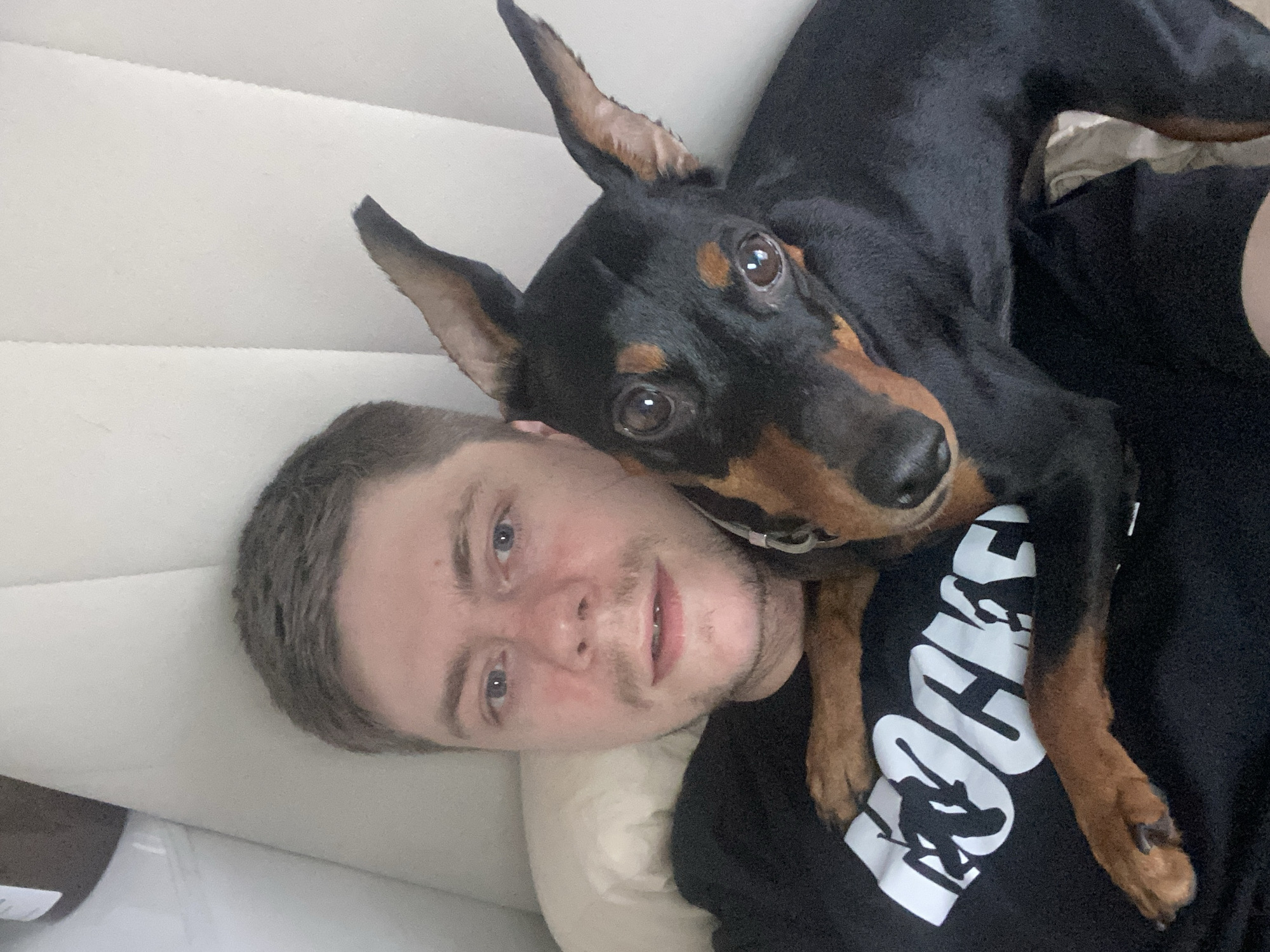 In a close-up, indoor selfie, a young man with short brown hair and blue eyes is pictured lying down, embracing a small black and tan dog. The man is wearing a black t-shirt with a white graphic design featuring a skateboarder. The dog, a Miniature Pinscher, rests its head on the man's chest, its paws draped over his shoulder and chest. The background consists of a light-colored, padded surface, likely a couch or bed, with a faint vertical seam. The lighting is soft and diffused. There is no discernible information to suggest this scene is located in Moscow, Russia.