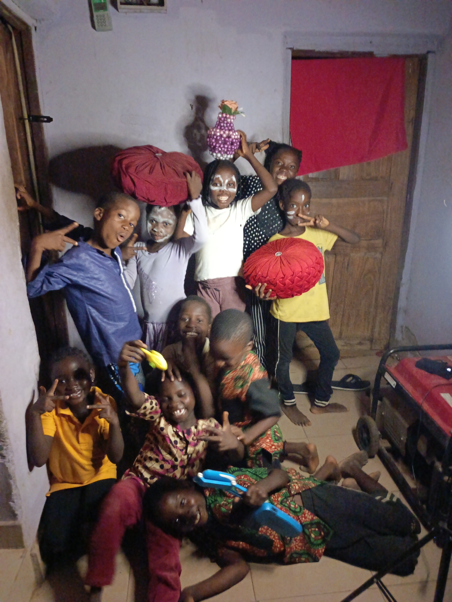 This is a vibrant indoor group photograph featuring approximately eleven children, all appearing to be of primary school age. The children are posing playfully for the camera, with many smiling widely, laughing, and making peace signs or other hand gestures. Several children have white face paint or powder applied in various patterns on their faces, suggesting a festive or celebratory occasion.
In the center, a child holds a red, round, ruffled cushion. Another child balances a purple beaded vase containing artificial flowers on their head. Other children are holding small yellow or blue plastic toys. The children are dressed in a variety of casual clothing, including t-shirts, dresses, and trousers, in different colors and patterns.
The scene is set indoors, likely in a room with light-colored walls and a tiled floor. Wooden doors are visible on both the left and right sides of the frame. A bright red piece of fabric is draped or hung in an opening in the upper right background. In the upper left, a wall-mounted intercom or phone device can be seen, but any text on it is unreadable due to distance and resolution. A red portable generator with wheels is partially visible in the bottom right corner of the image. The lighting appears somewhat dim, contributing to the intimate indoor setting. The overall mood conveyed by the children's expressions and poses is one of joy, playfulness, and camaraderie.