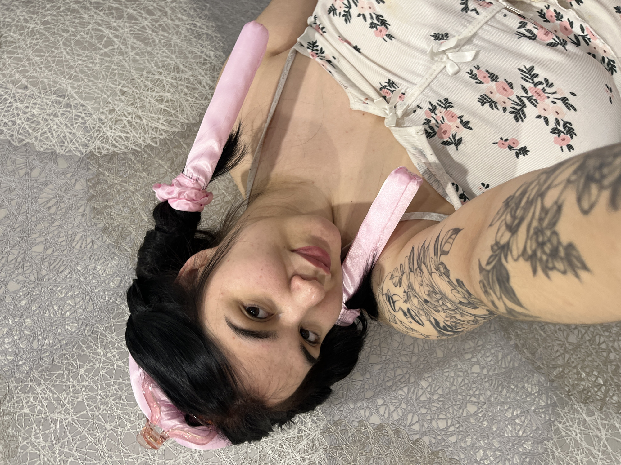 A woman is lying on a bed with a textured gray cover. She is wearing a white floral top with bows. Her dark hair is styled with pink rollers. She has a tattoo on her upper arm. The location is unknown, not specified in the description.