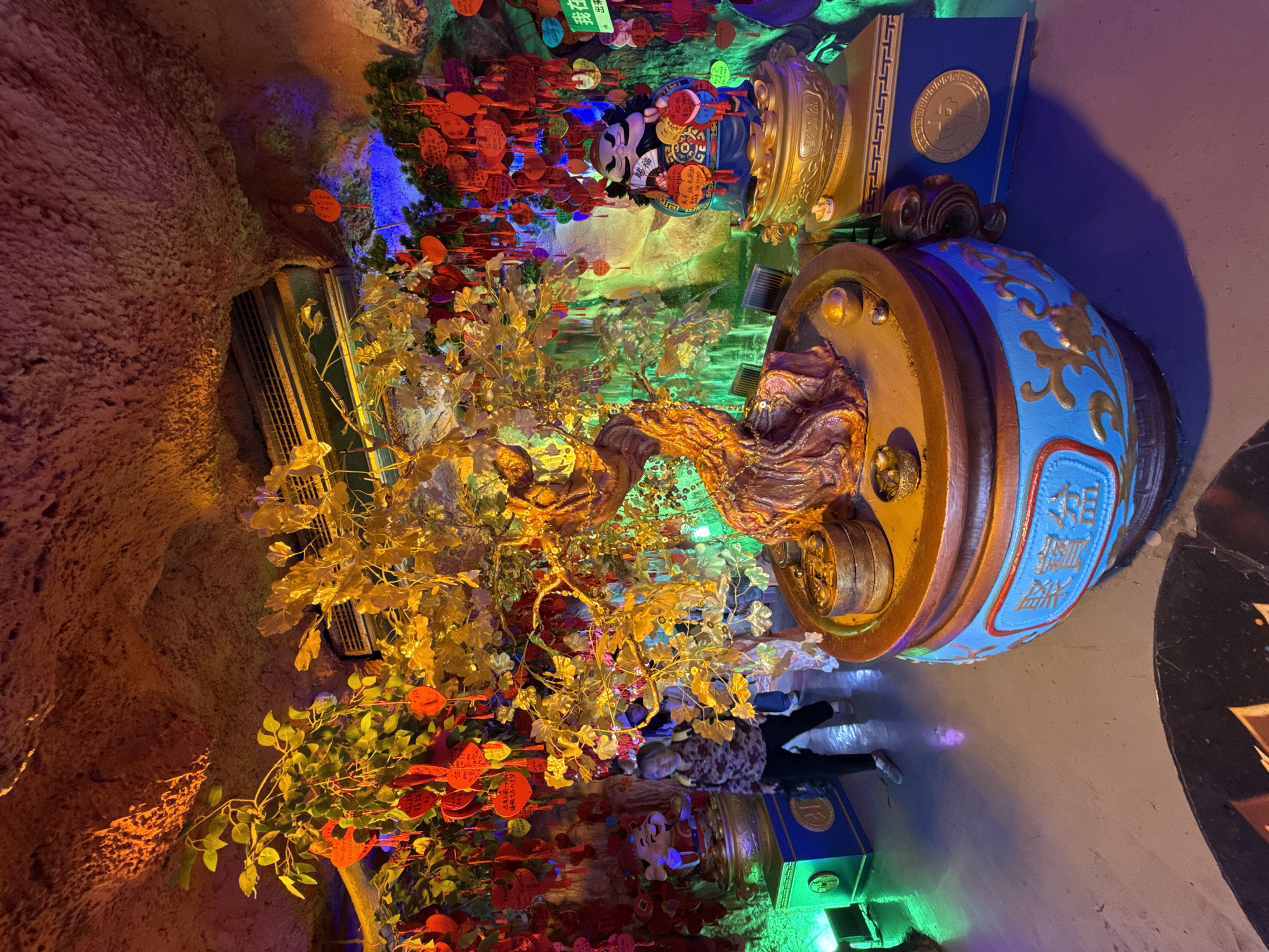 An interior shot, rotated 90 degrees clockwise, captures a vibrant, richly decorated space, likely a temple or cultural exhibit in Yuzhong District, China. The scene is illuminated by dramatic, colorful lighting in shades of blue, green, orange, and red, creating a mystical and festive atmosphere.
The central focus is a large, ornate "money tree" or "fortune tree" with golden, shimmering leaves, possibly artificial, emerging from a substantial, decorative golden pot. This pot features intricate blue and gold patterns, with visible Chinese characters that read "招财进宝" (zhāo cái jìn bǎo), meaning "ushering in wealth and prosperity." To the right (top when correctly oriented), another large golden vessel or statue is visible, adorned with similar cultural motifs.
On the left side of the frame (bottom when correctly oriented), another plant with green leaves is densely covered with numerous red, heart-shaped, and round paper charms or tags. Some of these tags also bear Chinese characters, including "福" (fú) for "fortune" or "blessing," and "我在" (wǒ zài) meaning "I am (here)." These charms suggest visitors leave wishes or dedications.
The background features textured, rocky, or cave-like walls, enhancing the immersive environment. A single person is partially visible near the bottom center (when correctly oriented), standing and observing the display. The activity suggests an interaction with cultural or spiritual offerings. Other decorative elements include small blue structures resembling books with gold medallions. The overall impression is one of cultural reverence and a wish for prosperity.