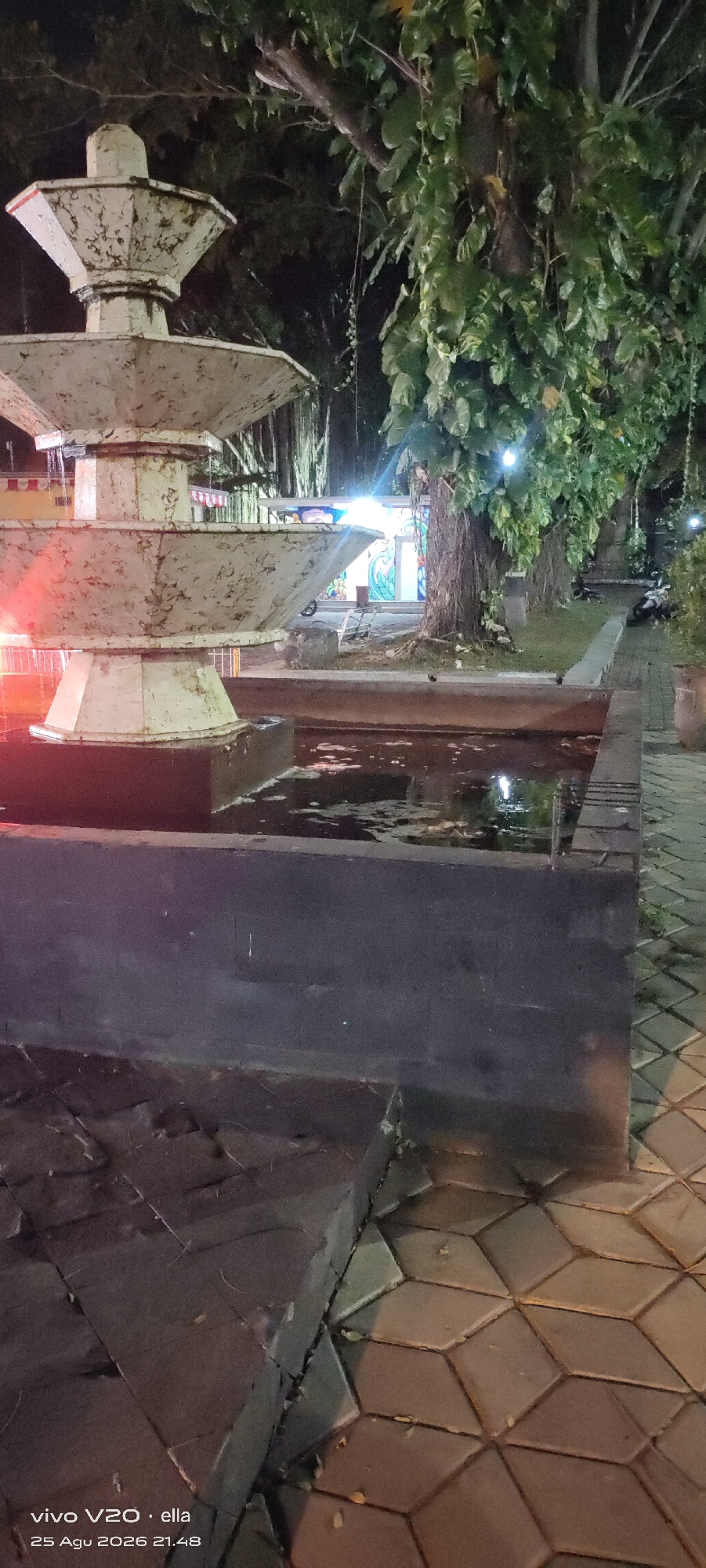 The image shows a tiered, white, weathered fountain at night, situated next to a pool of water. The fountain has multiple levels with water cascading down. In the background, there is a large tree with lush green foliage and visible roots. Several lights illuminate the scene, including what appear to be streetlights or decorative lights, casting a warm glow. A paved walkway with a diamond pattern is visible in the foreground and extends to the right. Part of a building with colorful murals is visible behind the fountain. There are some motorcycles parked in the distance. The overall impression is a public outdoor space at night, possibly a park or a public square. The timestamp at the bottom of the image indicates it was taken on August 25, 2026, at 21:48 in Tulungagung, Indonesia.