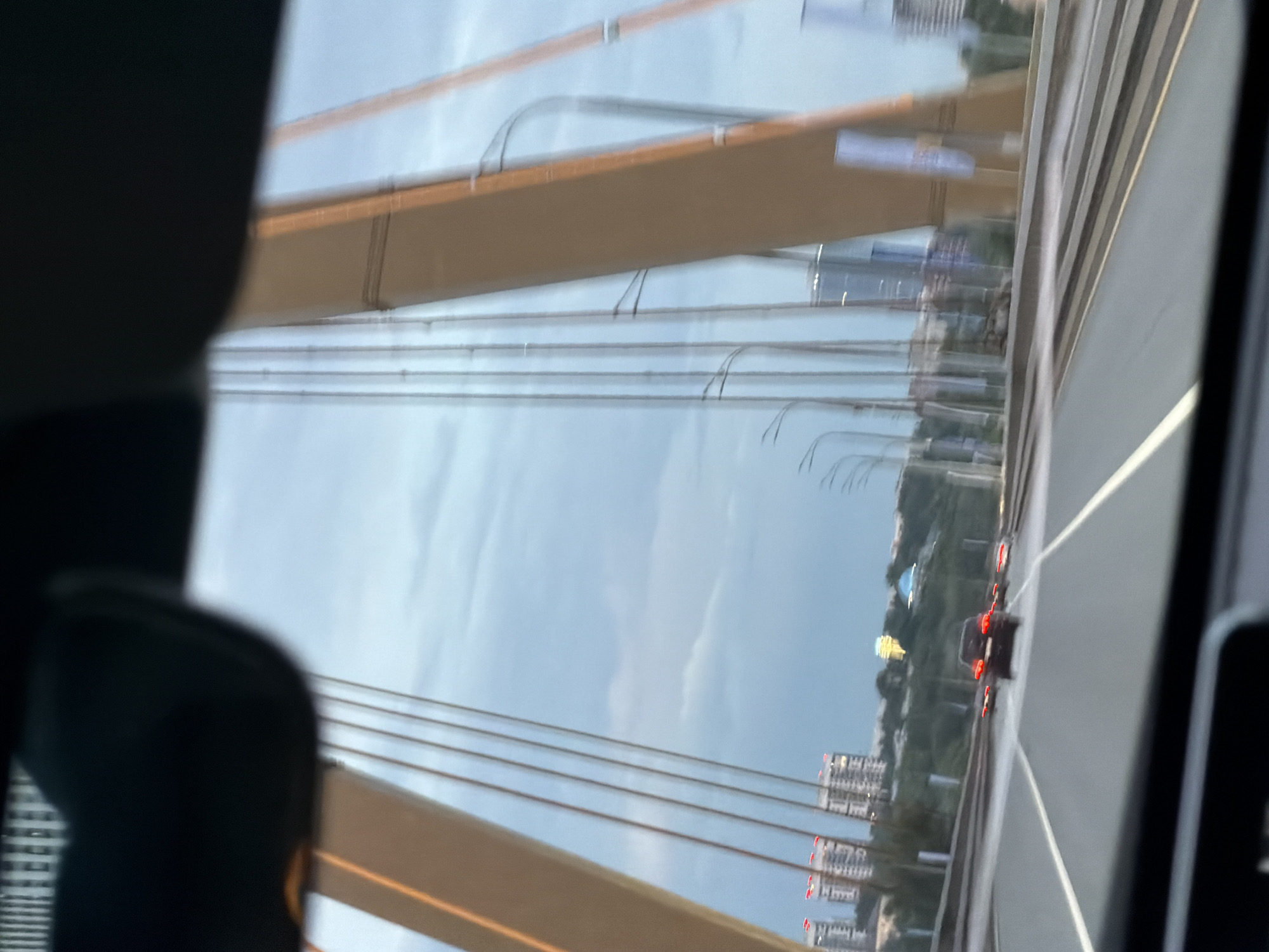 The image depicts a view from inside a vehicle traveling on a bridge, likely the Millennium Bridge in Kazan, Russia, given the distinctive architecture and urban landscape. The sky is overcast with a pale blue and grey hue. Tall, golden-brown pylons of the bridge are visible, crisscrossed by numerous cables.
In the foreground, the dashboard and windshield of the vehicle are visible, framing the scene. The road ahead is paved and marked with white lines, indicating multiple lanes of traffic. Several cars are driving on the bridge, with their taillights illuminated. The closest vehicle in the lane ahead is a dark-colored sedan.
In the background, a cityscape with several multi-story buildings can be seen on the horizon. Some of these buildings have red lights on top, possibly indicating their height. A prominent, glowing golden dome-like structure is visible in the distance, which is a characteristic feature of Kazan's skyline. The overall scene suggests a moment during twilight or a cloudy day, with a sense of movement and transit.