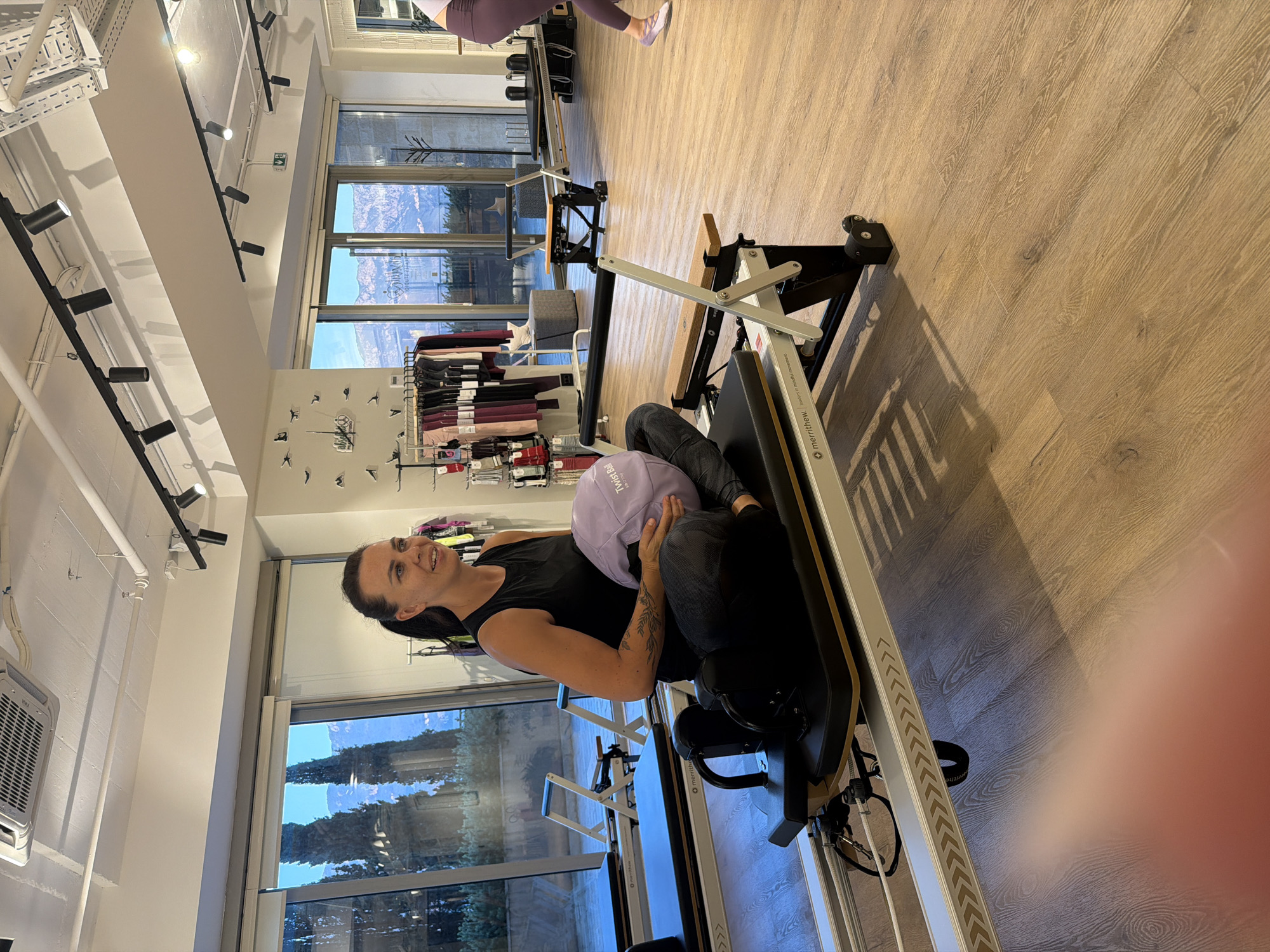 The image depicts a woman sitting on a Pilates reformer machine in a brightly lit studio. The woman is wearing a black tank top and patterned leggings, holding a light purple ball in her lap. Her hair is pulled back in a ponytail, and she is looking to her right with a slight smile.
The studio has a light wood floor and large windows that offer a view of a mountainous landscape with cypress trees. Several other Pilates reformers are visible in the background, as well as racks of exercise apparel. The room is equipped with modern track lighting and an air conditioning unit on the ceiling. The overall impression is that of a well-appointed fitness studio. The location is identified as Boreti, Montenegro.