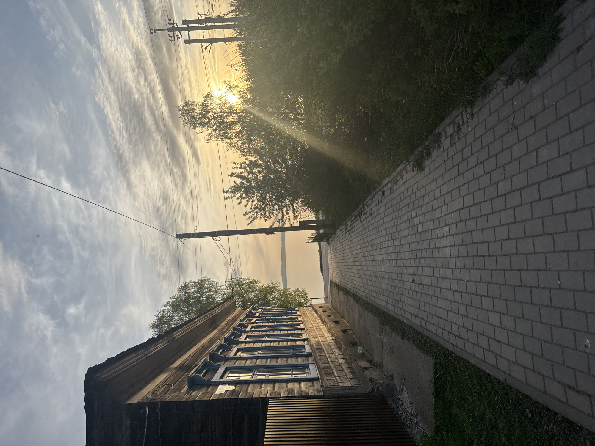 The image captures a rural scene near Osa, Russia, featuring a weathered wooden house with blue-framed windows set against a sky washed with the golden hues of sunset or sunrise. Overhead power lines stretch across the sky, and the sun filters through the trees, casting rays onto the scene. A brick-paved pathway extends from the house into the distance. Lush greenery borders the path and surrounds the structures, blending with the natural landscape. The angle of the shot, which could be taken by a person standing, causes an illusion of the building pointing up. The buildings are a reflection of the old architecture of Osa. The view is of the Kama River.