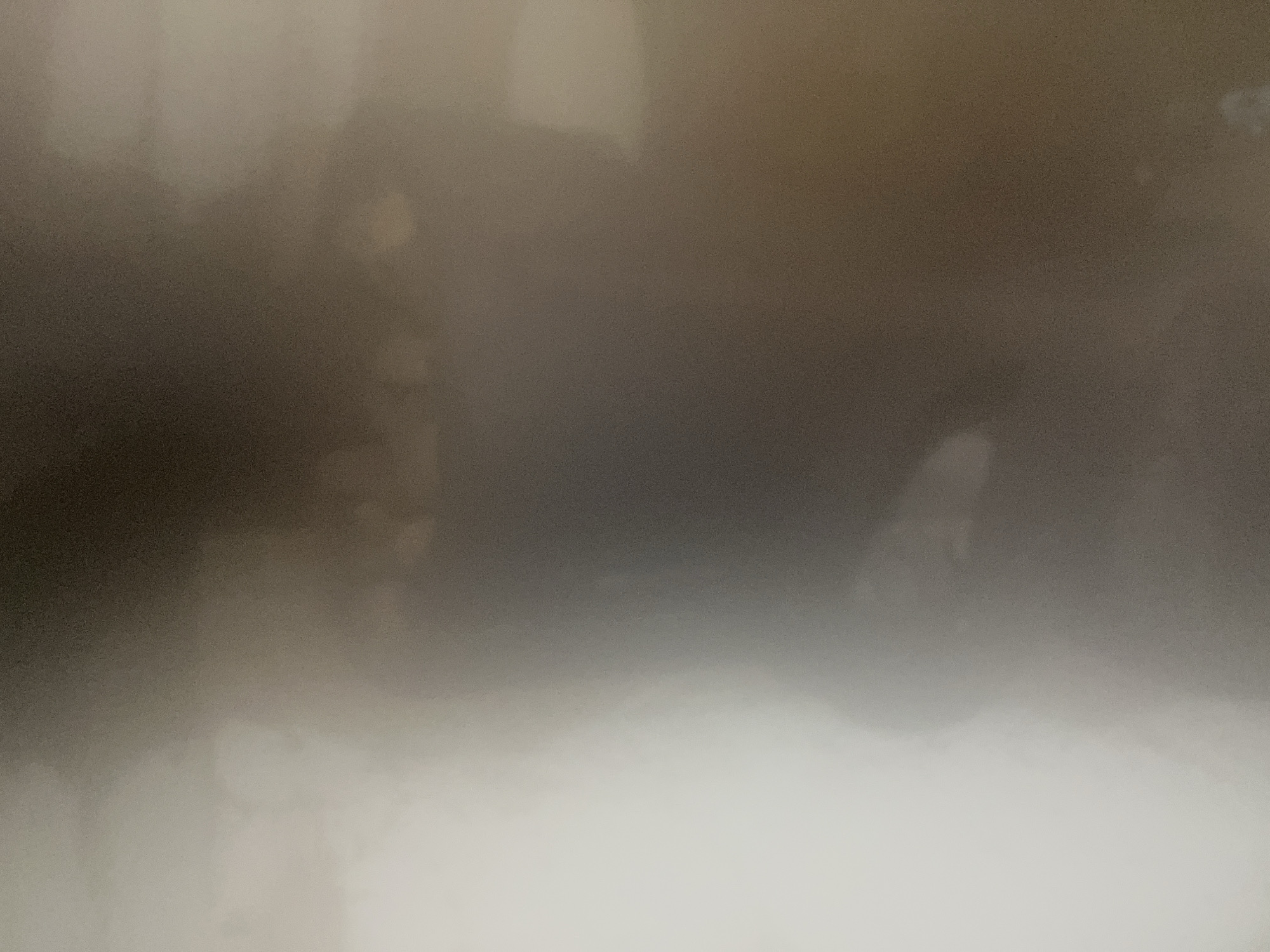 This is a close-up, out-of-focus photograph of a person's torso. The subject is wearing a dark-colored shirt. The lighting is dim and diffused, with a bright light source to the left of the frame, creating a gradient of light and shadow across the image. The background is indistinct, appearing as a mix of dark and lighter shades. The image lacks any clear environmental context, and therefore, its location within the city of Уральское, Russia, cannot be determined.