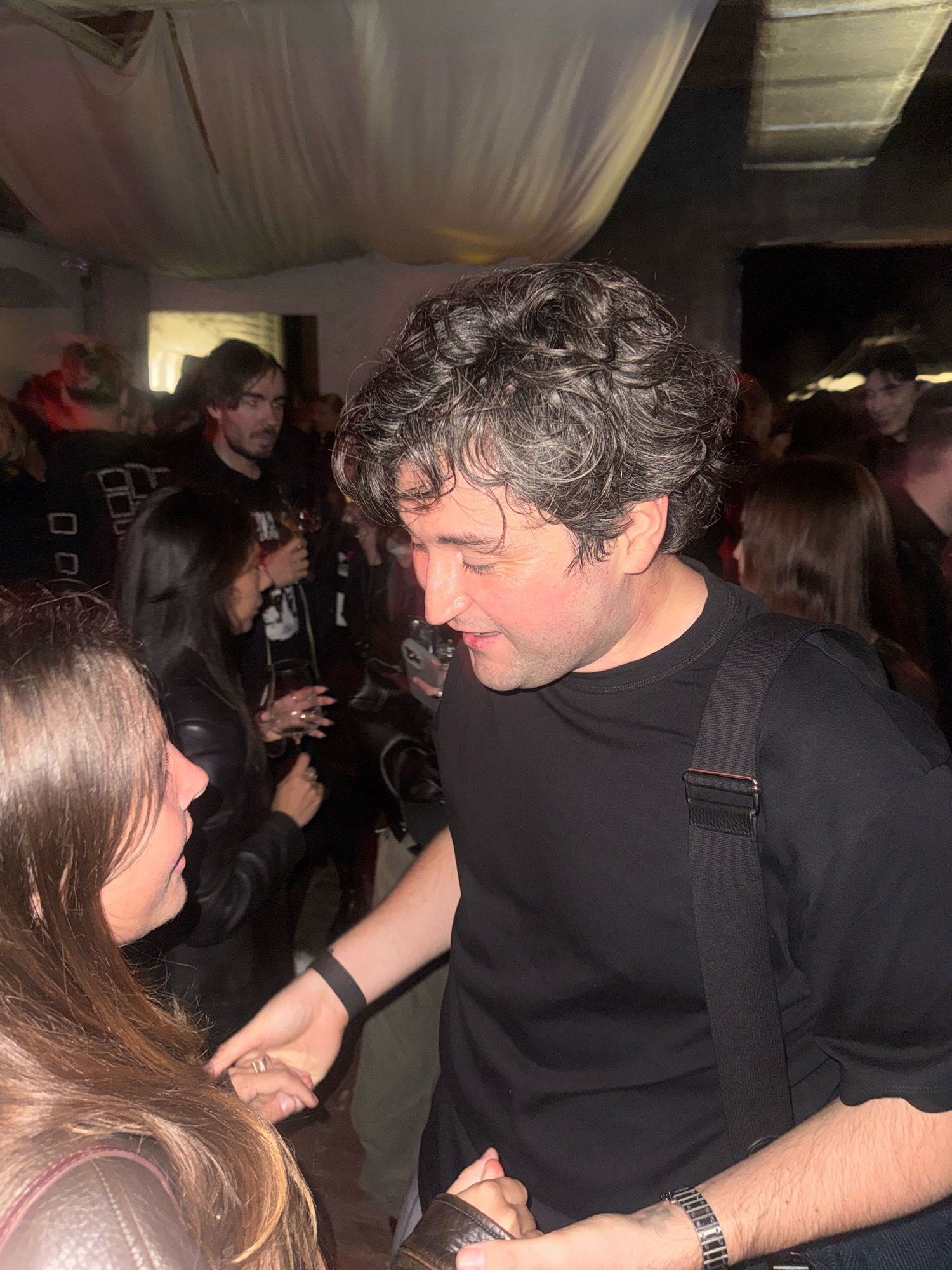 The image depicts an indoor gathering, possibly a party or a bar, with several individuals present. A man with curly graying hair and a black t-shirt is in the foreground, looking down and to his left. He is wearing a black bag slung across his body and a watch on his left wrist. To his left, a woman with long brown hair is partially visible, looking towards the man. Their hands are touching, suggesting an interaction. Other people are visible in the background, some holding drinks. The environment appears dimly lit, with fabric draped from the ceiling, creating a textured backdrop. The location is not specified as being in Moscow, Russia.