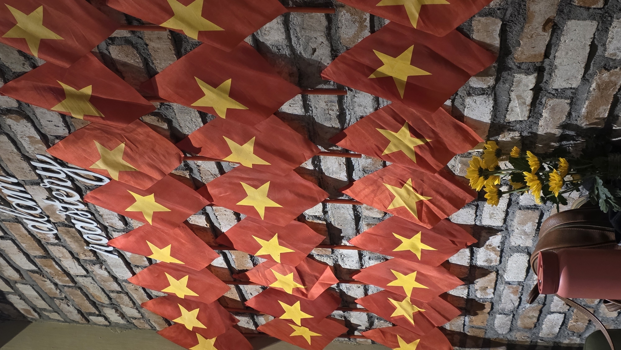 The image depicts a wall decorated with numerous small Vietnamese flags. The wall appears to be made of weathered bricks. To the right of the flags, there is a bouquet of yellow flowers and a pink water bottle resting on what seems to be a backpack. Additionally, a partial sign with the words "roastery" is visible on the left side of the image. Given the prevalence of the Vietnamese flags and the text "roastery," it is possible that the location is a coffee shop in Nha Trang, Vietnam, a city known for its coffee culture.