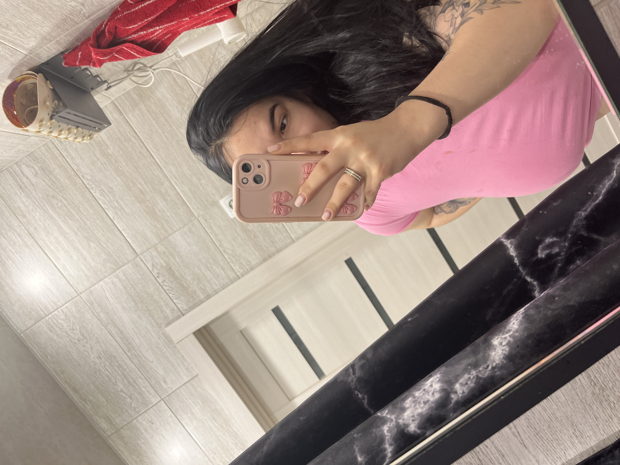 The image is a mirror selfie taken by a female individual within a room in Chaikovsky, Russia. The individual has long dark hair and wears a pink t-shirt. Her right hand, adorned with a multi-banded ring on the ring finger and light pink nail polish, holds a light pink iPhone in a case decorated with bows, obscuring the lower portion of her face. Her left arm, visible on the right side of the frame, displays a black elastic hair tie on the wrist and a floral-patterned tattoo on the upper arm. The background, reflected in the mirror, features walls tiled with light-colored, wood-grain patterned tiles. A light-colored interior door with two dark horizontal panels is partially visible on the left. On the upper right, a dark gray wall-mounted shelf holds a beige, perforated, rolled-up bath mat. A red and white striped towel hangs from a hook below this shelf, next to a white electrical cord and a cylindrical white device mounted on the wall. A dark surface with a white marble-like pattern is visible in the immediate foreground, consistent with a shelf or the mirror's lower frame.