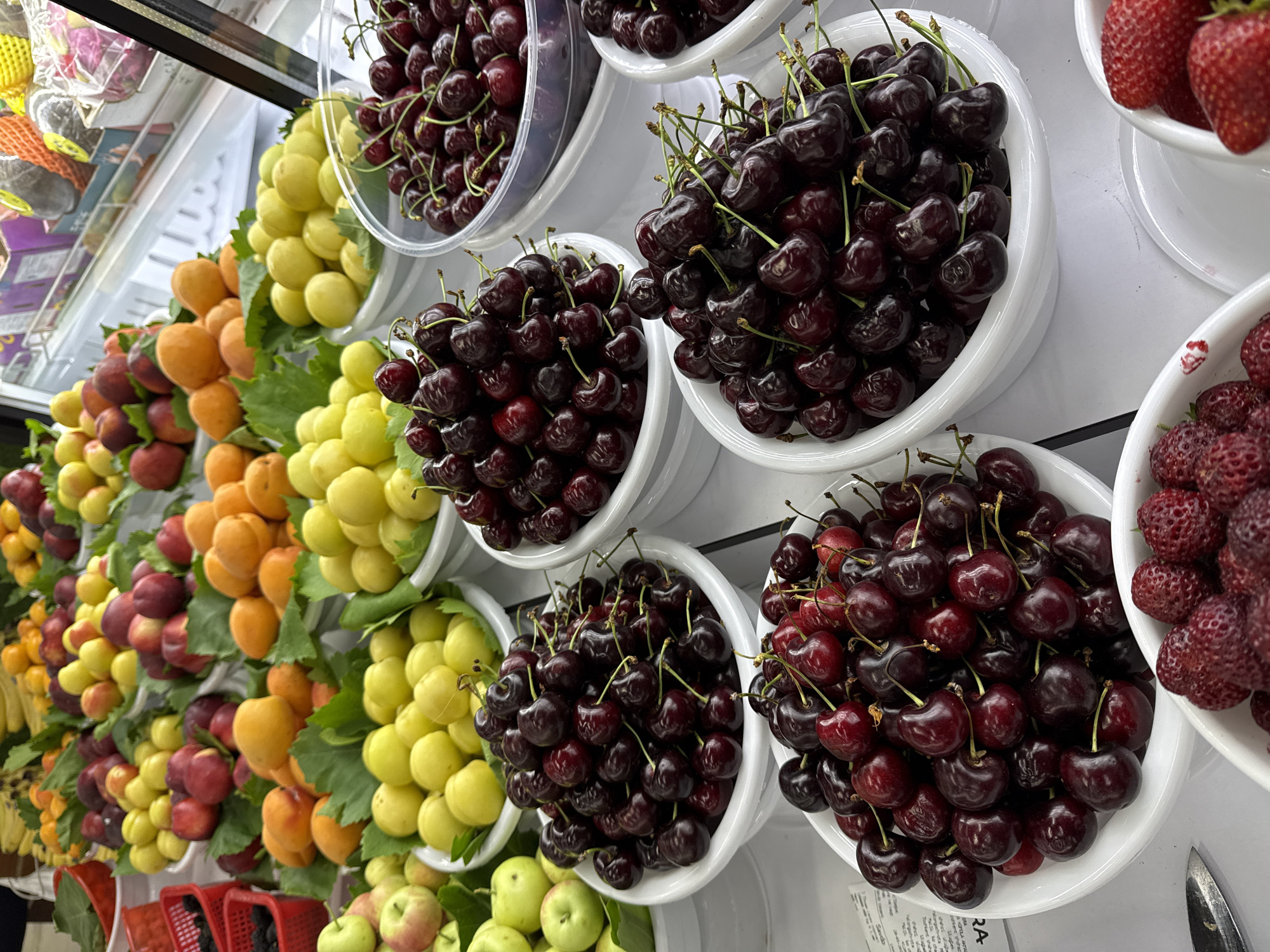 The image depicts a fruit display, likely inside a grocery store or market in Tashkent, Uzbekistan. Multiple white bowls are stacked and filled with different fruits: dark red cherries, bright red strawberries, yellow plums, red plums, orange apricots, and green apples. A clear plastic container is visible at the top of the frame also containing cherries. The fruits appear to be fresh and well-stocked. In the lower left, red shopping baskets are visible. The background is generally out of focus, but shelving and packaged goods are discernible.