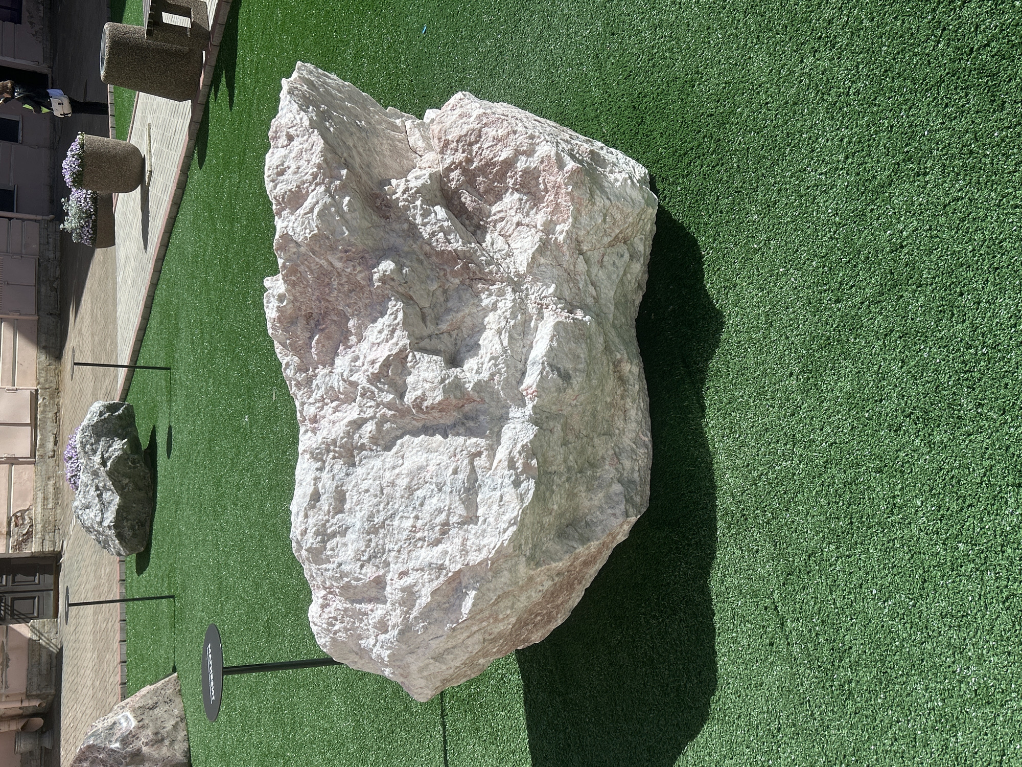 An outdoor scene features a large, light-colored boulder with pinkish veins, resting on artificial green turf. The boulder dominates the foreground, casting a shadow to its left. In the middle ground, another, larger gray boulder with a cluster of purple flowers on top sits on the turf. Behind this, a paved area with a brick border is visible, with two large, stone planters filled with purple flowers. To the right, a cylindrical concrete object with a metal bench is present. In the far background, a building with pinkish walls and a dark doorway is visible, with a person in dark clothing carrying a white bag walking away from the scene on the paved area. A black, circular sign with white lettering is mounted on a pole in the middle ground. The lighting suggests a sunny day.