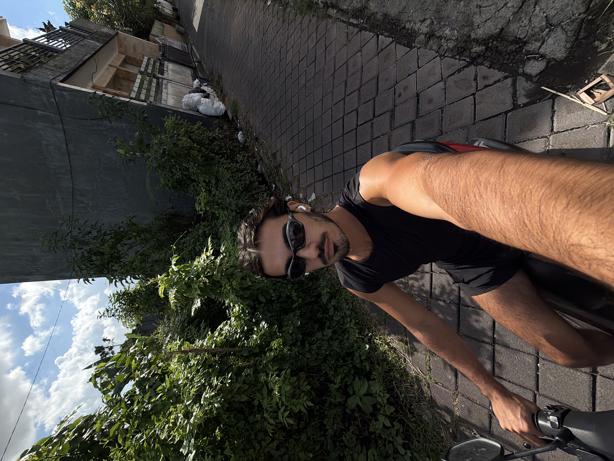 The image shows a man riding a motorcycle on a narrow, brick-paved street. The man is wearing sunglasses and dark clothing. He appears to be taking a selfie while riding. Lush greenery borders the road on one side. The other side has a building, with some trash bags visible. The sky is visible with some clouds. The location is identified as North Kuta, Indonesia, based on contextual elements.