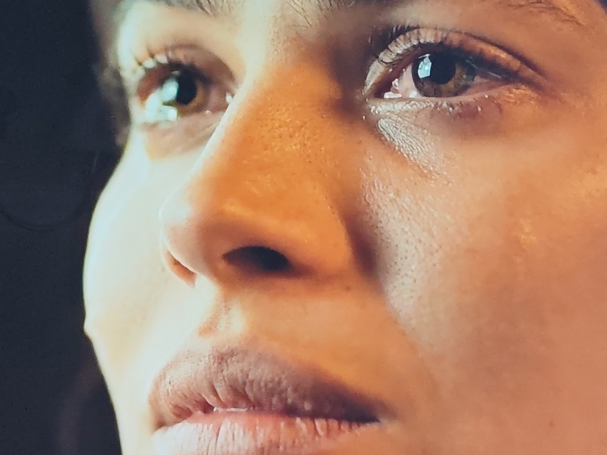 The image is an extreme close-up of a person's face, focusing on the eye, nose, and mouth. The lighting is warm and highlights the texture of the skin, including pores and some blemishes. The subject's eye is brown with visible eyelashes. There appears to be some white residue around the eye, possibly makeup or a residue from tears. The nose is centrally located in the frame and appears to be a light brown or tan. The mouth is slightly open, revealing teeth and a pink tongue. The lips are full and detailed with visible texture. The background is dark and out of focus, providing no contextual information about the location or environment. There is no indication of the city or any specific location.