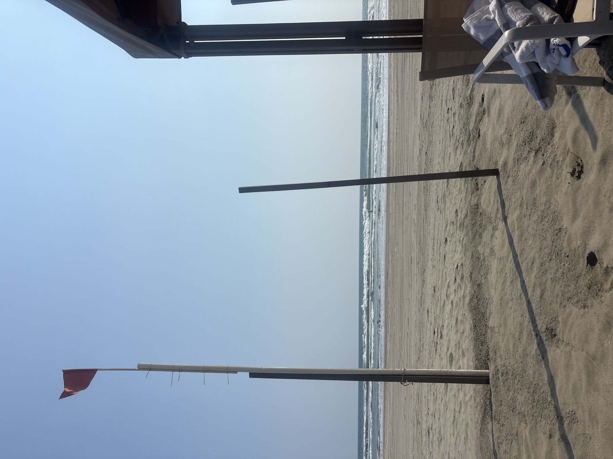 The image depicts a beach scene in Acapulco de Juárez, Mexico, under a clear, bright blue sky. In the left foreground, a tall, light-colored pole, possibly wood, is anchored in the sand. A red rectangular flag is affixed to its top, unfurled horizontally to the right, indicating wind presence. Several smaller horizontal protrusions are spaced along the pole's left side. A rope is tied near its base. To the right of this, a shorter, darker vertical pole is also embedded in the sand, casting a distinct shadow towards the right. In the far right foreground, a partial view of a multi-post structure, likely a beach cabana or shelter, is visible. Beneath this structure, a light-colored beach chair holds two folded towels: one predominantly white with blue accents, and another striped gray and white. The beach consists of light brown sand, showing numerous parallel tracks, consistent with vehicle activity, and scattered footprints. In the midground, the ocean extends to the horizon, featuring multiple breaking waves. A small, dark, humanoid figure is visible in the water beyond the immediate wave line. No other prominent individuals are present. Strong sunlight is indicated by the clear shadows cast by the poles.
